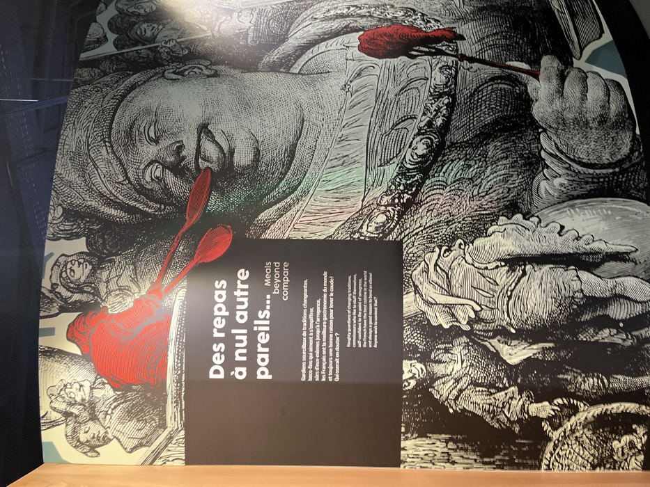 This image displays a large, brightly lit exhibition panel located within the Cité internationale de la gastronomie et du vin in Dijon, France. The panel features an intricate, black-and-white illustration in an engraving style, with specific elements highlighted in red.
The central subject on the right is an extremely corpulent man depicted in profile, facing left. He wears a large, elaborate turban and an ornate, heavy-linked necklace or collar. His eyes are rolled back, and his tongue extends from his mouth, suggesting intense sensory pleasure or gluttony. He holds a thin, long-handled utensil, possibly a spoon or small fork, with a vibrant red, dripping substance at its tip, positioned near his mouth. His left hand, largely obscured, appears to hold a bowl or container from which the substance was taken.
In the upper left quadrant, a group of smaller, less defined figures observe the scene. One figure, centrally positioned within this group, is entirely highlighted in red and appears to be offering or presenting two red spoons.
The lower left section of the panel shows two additional figures, also in the black-and-white engraving style. One stands upright, looking right, while the other, with its back partially to the viewer, is a grotesque or heavily cloaked figure holding a large, rounded object resembling a pot or barrel.
Overlaid on the illustration, primarily in the left and center, is white text. The most prominent title reads "Des repas à nul autre pareils..." followed by "Meals beyond compare" in English. Below this, a French paragraph describes "Haughty guardians of changing traditions, connoisseurs who like to stuff themselves... the French have the best cuisine in the world and always a good reason to bend an elbow!" with an accompanying English translation. The overall theme conveyed is a celebration of French gastronomy, abundance, and culinary pride.