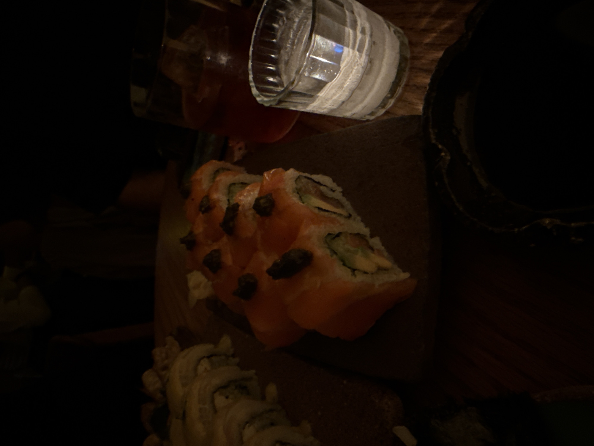 The image shows a close-up view of a plate of sushi, a glass of liquid (possibly a cocktail), and another bowl-like object, likely in a dim or dark environment. The sushi appears to be a salmon roll with dark toppings, possibly caviar or a similar ingredient. A second type of sushi is visible in the lower portion of the image. The glass contains ice. The scene is possibly in a restaurant or bar setting. Without additional information, determining the specific location in Mexico City is not possible.