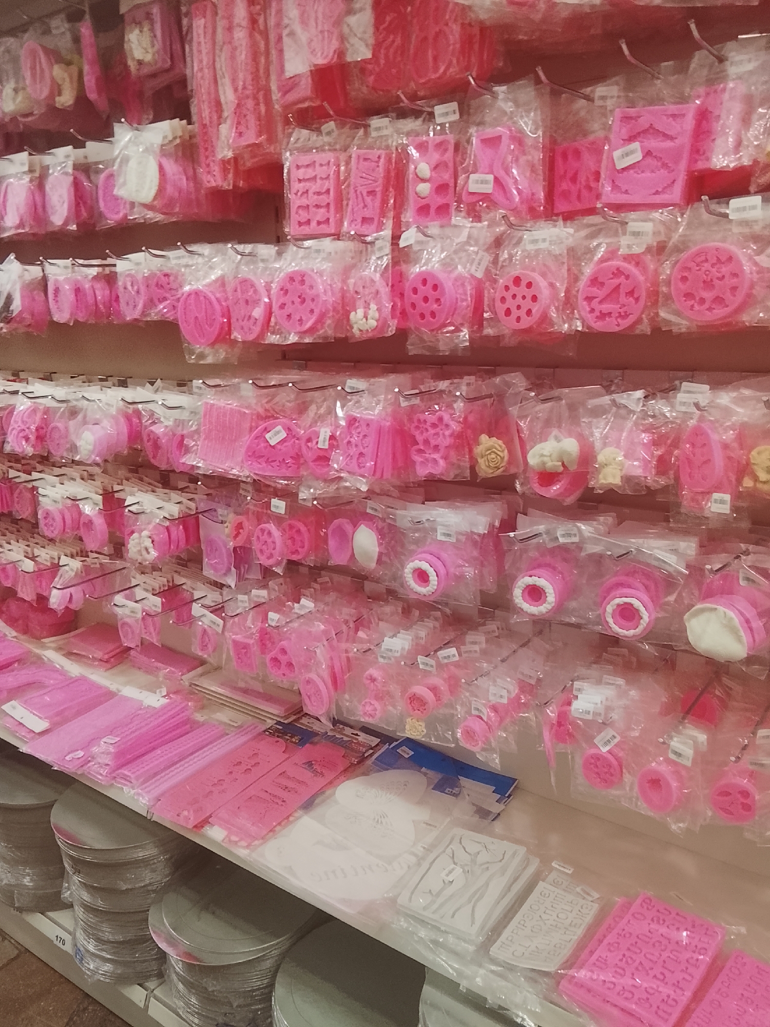 A detailed view of an organized retail display within a store shows multiple shelves stocked predominantly with pink silicone molds for baking and crafting. The items are individually packaged in clear plastic and hung on metal hooks attached to a slatwall or pegboard system on the back of the shelves.
The top shelves feature a variety of molds, including those for letters (such as "LOVE" and individual numbers), intricate patterns, and floral designs. Some packages contain small, finished white or cream-colored molded shapes, indicating the use of the products.
The middle shelves continue with a wide assortment of pink silicone molds, featuring circular designs with various cutouts (e.g., dots, heart shapes, a number '4'), star shapes, leaf patterns, and heart designs. Also present are cylindrical molds, some featuring an inner white ring, suggesting multi-part designs.
The lowest visible shelf displays stacks of flat pink items, possibly textured mats or additional molds, laid directly on the shelf. Adjacent to these are several white stencils or mats with cut-out designs, including a deer head and various alphabets or patterns.
Below the shelving units, on a lower rack or floor, are multiple stacks of round, silver-colored discs, likely cake boards or bases, wrapped in plastic. One stack has a white label with "170" printed on it.
Each packaged item generally bears a small white barcode or price label. The overall color scheme is dominated by various shades of pink, with accents of white, cream, and silver. The lighting is even and artificial, characteristic of an indoor retail setting. No people are visible in the image.
