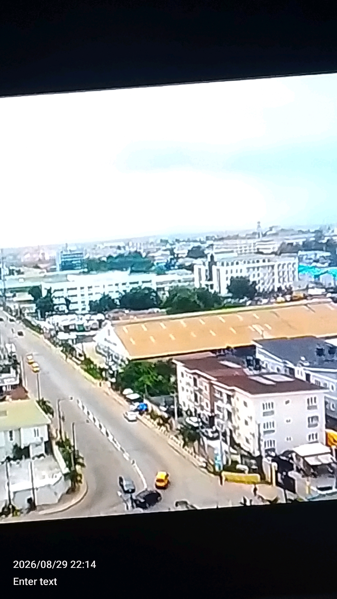 The image is an aerial view of a street scene in Lokoja, Nigeria. A paved road curves through the urban landscape, with several vehicles visible. On the left side of the road, there are white and light-colored buildings with multiple stories and visible windows. Further into the background, there are larger industrial-style buildings with long, tan-colored roofs, surrounded by trees. The sky is overcast. In the foreground, at the bottom of the image, text indicating a date and time "2026/08/29 22:14" and "Enter text" is displayed.
