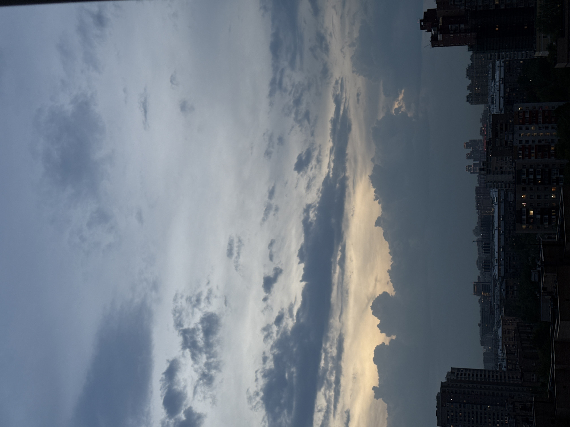 The image depicts a cityscape at dusk, likely Moscow, Russia, given the architecture. In the foreground, a cluster of high-rise buildings is visible, some with illuminated windows. In the background, the sky is filled with dark, gray clouds, punctuated by patches of lighter gray and hints of sunlight. The light suggests a fading sunset. The scene appears to be captured from an elevated vantage point, possibly a high-rise building.