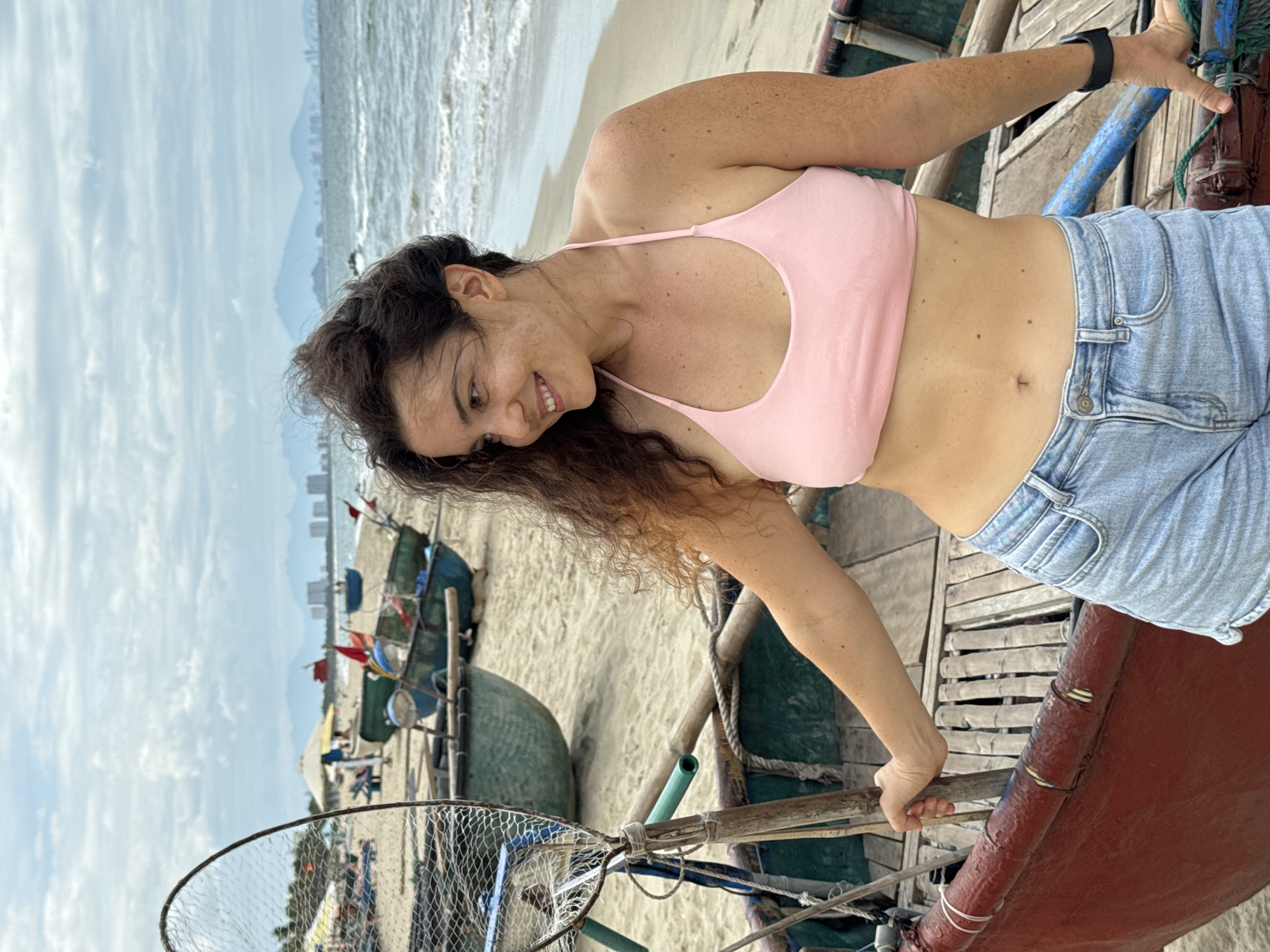 An outdoor, medium shot captures a young adult woman posing with a smile next to a round boat on a beach in Hoi An, Vietnam. The woman, who has curly brown hair and is looking to her right, is wearing a pink bikini top and light blue denim shorts. Her left hand rests on the edge of the boat, and her right arm is bent at the elbow. The boat, predominantly red with green interior, is wooden and appears to be a traditional fishing vessel. Behind the woman and boat, there are other similar boats on the sand, some with fishing nets. The ocean is visible in the background, with small waves gently rolling onto the shore. In the far distance, a city skyline with buildings and mountains can be seen under a cloudy sky. The beach is sandy, and there are beach umbrellas and lounge chairs scattered around. The scene suggests a casual, relaxed atmosphere, typical of a coastal tourist destination.
