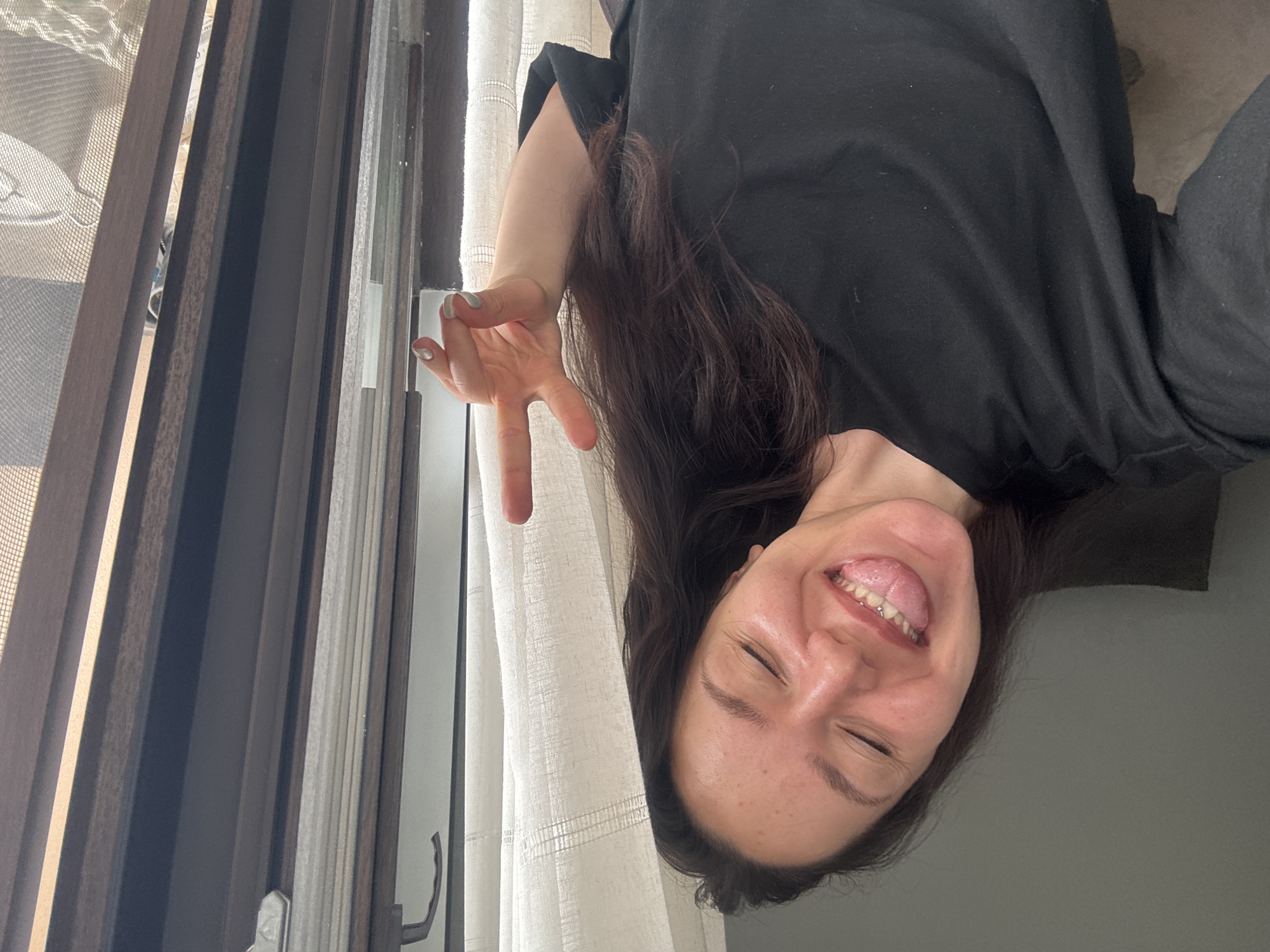 A woman is shown in a selfie. She's sticking her tongue out and making a hand gesture. She has dark hair and is wearing a black shirt. Behind her is a window with a curtain and mosquito net. The location cannot be determined based on visual information.