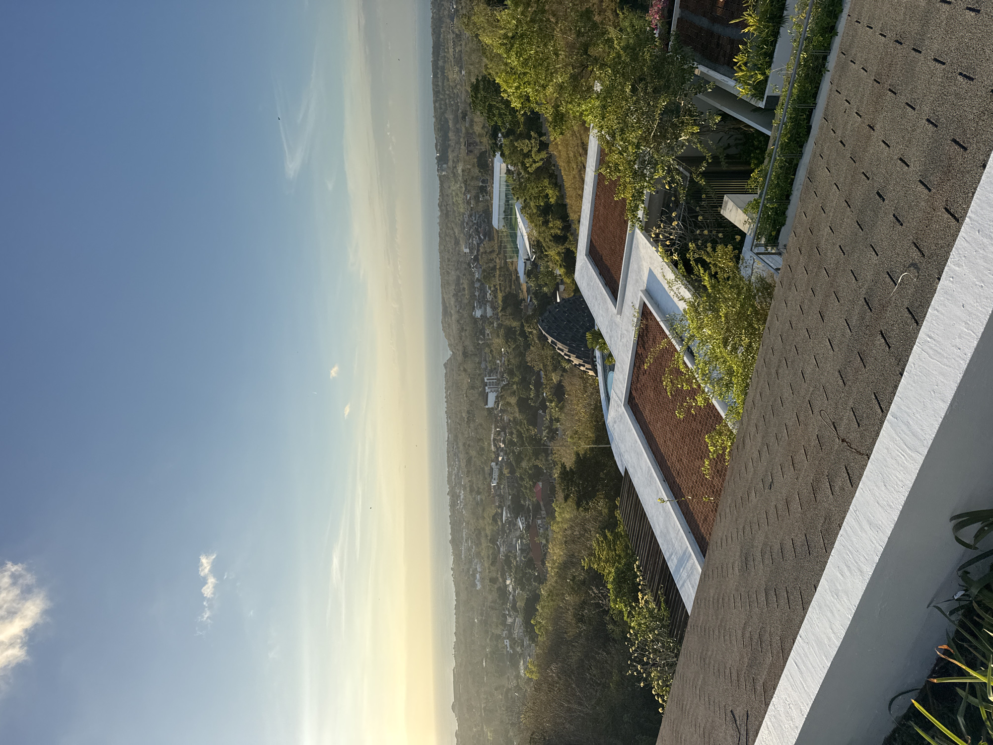 This image shows a high-angle view of a coastal city, likely Kuta Selatan, Indonesia, at sunset. The sky is a clear blue with a few wispy clouds, and the setting sun casts a warm, golden glow over the horizon and the ocean. Below the sky is a sprawling cityscape, characterized by numerous buildings, lush green trees, and what appears to be a large white structure with a green field, possibly a sports complex. In the foreground, the roofs of several modern buildings are visible, featuring a combination of dark shingles and reddish-brown tiles, interspersed with white architectural elements. One building has a distinctive angular, dark-tiled roofline. The overall impression is one of a developed, tropical urban environment bathed in the soft light of dusk.
