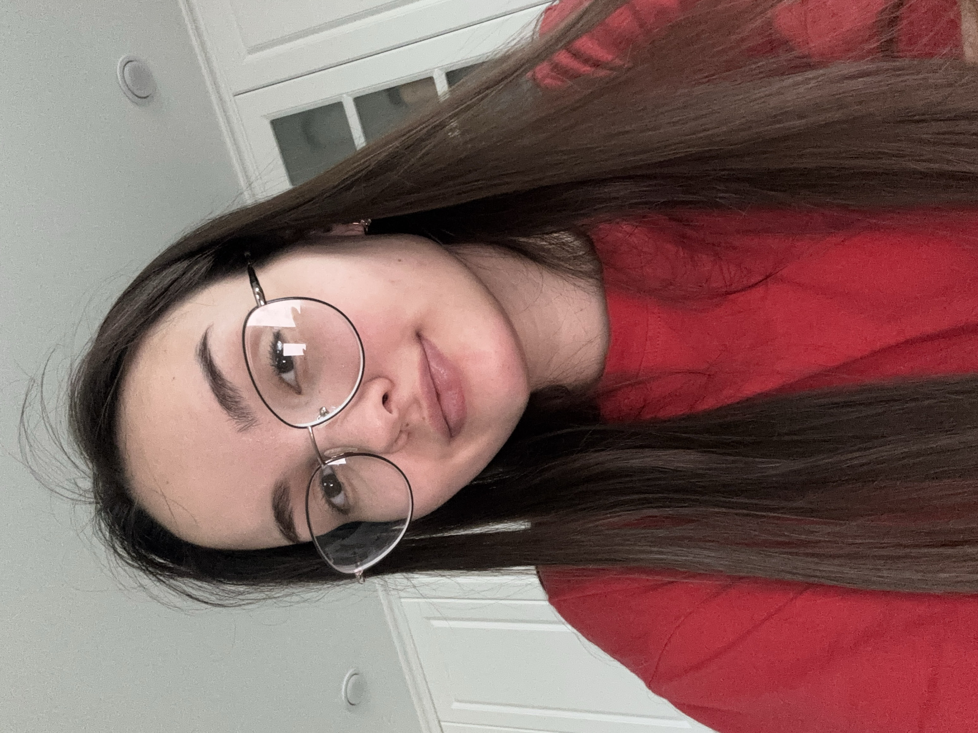 A young woman with long brown hair is wearing round, wire-framed glasses and a red shirt. The background is a plain white wall and a white paneled door. The location is not discernible from the image.