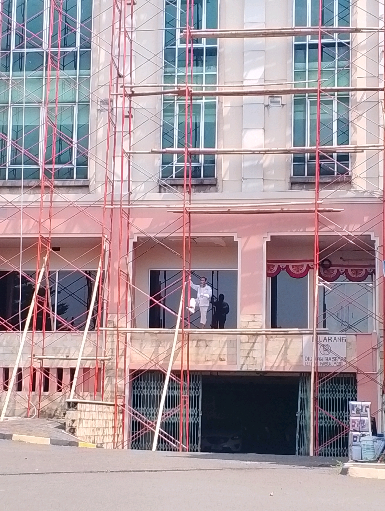 The image shows the exterior of a building undergoing renovation, indicated by the presence of scaffolding. The building appears to be multi-story, with large windows. On the second floor, a person in white clothing is visible standing on a balcony. Below this level, there is a dark opening, possibly an entrance to a basement or garage, covered by a metal gate. A sign above this opening reads "DILARANG" and "DIDALAM BASEMENT KELUAR MASUK MOBIL". The scene is set in a city environment with a paved road in the foreground.