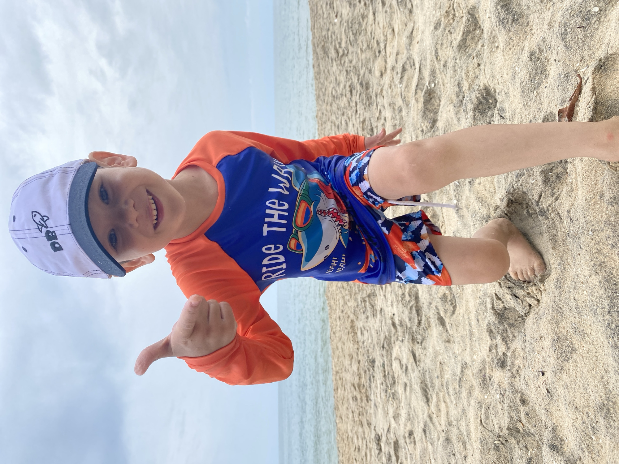 A young child wearing a white hat, orange long-sleeve shirt, and blue swim trunks kneels on a sandy beach. The child smiles and gives a thumbs-up gesture. The background shows a calm ocean meeting the sky, both obscured by light cloud cover. The scene is located on a beach in Phu Thuy Ward, Vietnam.