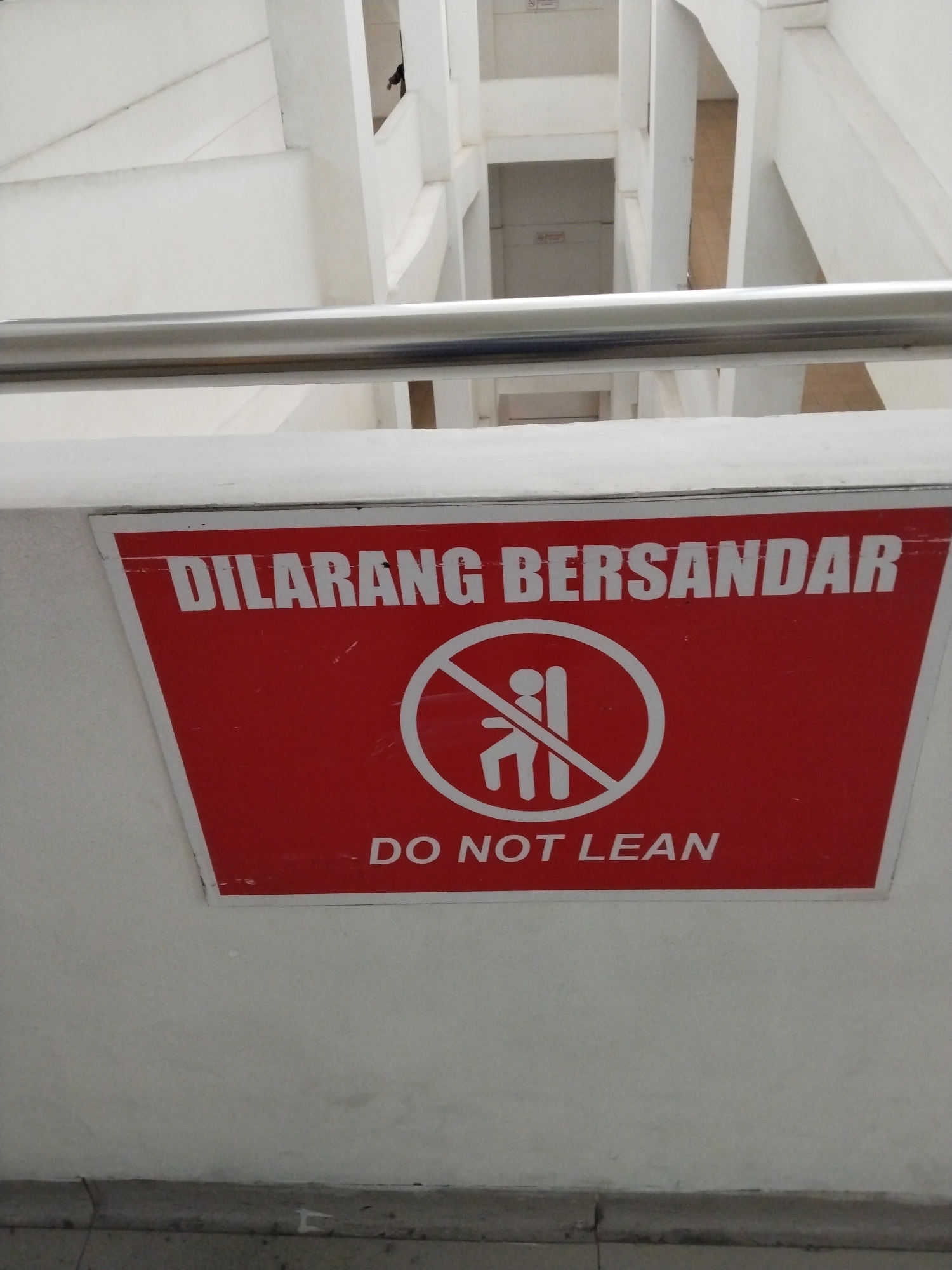 A red and white sign is mounted on a white wall, warning people not to lean. The sign features white text in Indonesian that reads "DILARANG BERSANDAR" and in English "DO NOT LEAN". Centered on the sign is a white circular graphic with a red diagonal line through it, superimposed over a pictogram of a person leaning against a railing. The background of the image shows a concrete stairwell with white railings. The lighting suggests an indoor environment, possibly a public building or infrastructure. There are no people clearly visible in the image, only the sign and the architectural elements of the stairwell. The image quality is moderate, with some minor scratches and debris on the sign. The location is not specified within the image itself, and no information about Bojonegoro, Indonesia is provided.