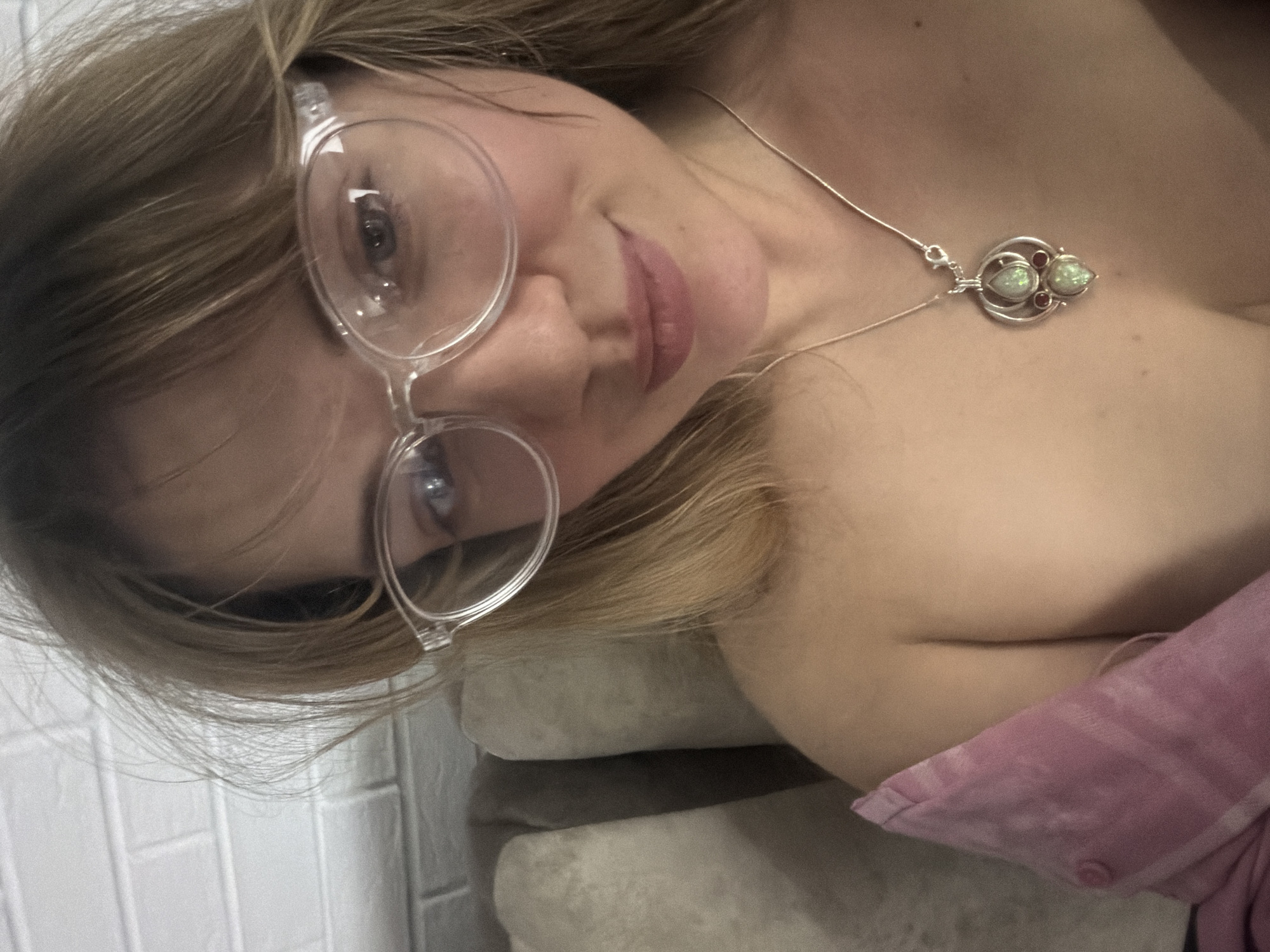 A close-up, slightly angled photograph captures a woman with light blonde hair and fair skin. She is wearing clear-framed glasses and a delicate silver necklace with a pendant featuring two pear-shaped, opalescent stones and two small, round, reddish stones. Her lips are visible and appear to be lightly tinted. She is reclined on what appears to be a soft, upholstered surface, with a portion of a pink garment or blanket visible in the lower right corner. In the background, to the left, is a white brick wall. There is no information to place the subject in the city of Большая Шильна, Russia.