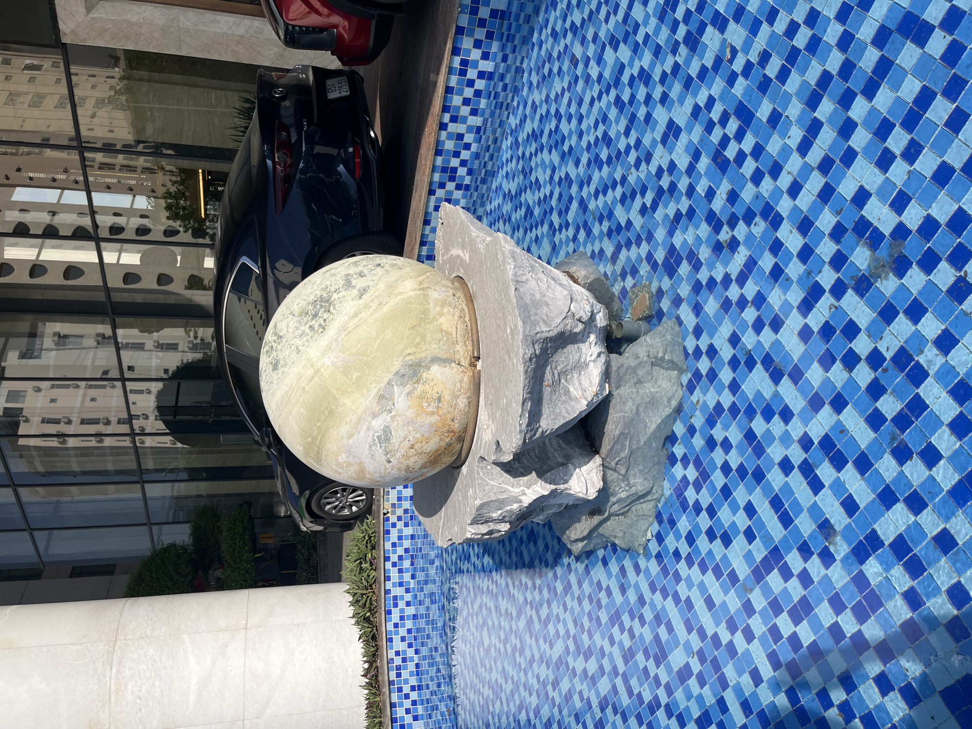 The image displays an outdoor water feature in what is identified as Bac Nha Trang Ward, Vietnam. In the foreground, a rectangular pool or fountain is tiled with a checkerboard pattern of dark blue and light blue mosaic squares. The water in the pool is clear and shows reflections of the sky and the stone structure within. Centered within this pool, a large, rough-hewn, grey rock formation serves as a base for a prominent spherical object. This sphere is mottled light green and yellow, with areas of weathered texture and discoloration.
Behind the water feature, a dark blue Mazda sedan (specifically, a Mazda 3) is parked, visible from its rear left quarter. The license plate "65A 275.51" is clearly legible. To the immediate right of the Mazda, the front-left section of a red vehicle, possibly an SUV or truck, is visible. Behind the cars and fountain, a modern building with a facade of large glass panels reflects the surrounding environment, including what appears to be another multi-story building with numerous windows and balconies. A large, smooth, white cylindrical pillar stands to the far left of the frame, partially obscuring the building. Green foliage, including shrubs and small trees, is visible between the cars and the reflective building, and along the edge of the fountain. The scene is illuminated by bright sunlight, casting distinct shadows. No individuals are present in the image.