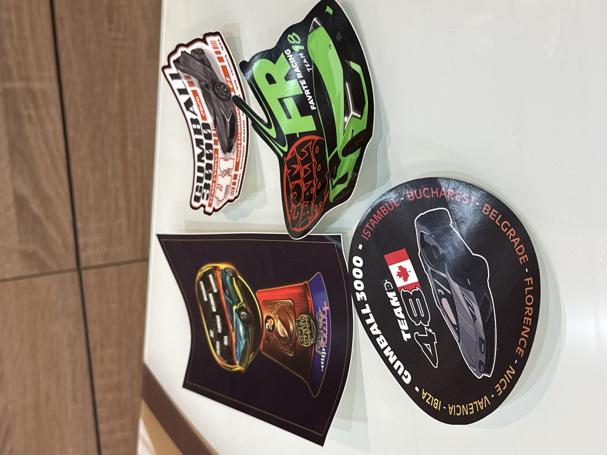 The image shows four stickers adhered to a white surface, likely a refrigerator or whiteboard. The stickers vary in shape and design, all appear to be related to the Gumball 3000 rally, a popular international car race. One sticker is circular and includes cities along the rally route, such as Istanbul, Bucharest, and Belgrade, along with the text "GUMBALL 3000" and "TEAM 48." Another sticker is shaped like a gumball machine. The third sticker is green and black with the text "FAVRTE RACING TEAM 18." The last sticker contains the words "Gumball 3000." There is no indication of a location in Prisovo, Bulgaria.