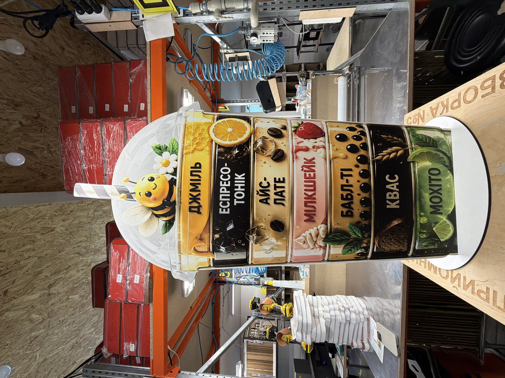The image shows a large, stylized cup of layered beverages, resembling a promotional display. The cup features distinct layers, each with corresponding text and imagery. From top to bottom, the layers include: "ДЖМІЛЬ" with a bee and honeycombs, "ЕСПРЕСО-ТОНІК" with a lemon slice, "АЙС-ЛАТЕ" with coffee beans and ice, "МІЛКШЕЙК" with whipped cream and a strawberry, "БАБЛ-ТІ" with tapioca pearls and leaves, "КВАС" with wheat, and "МОХІТО" with lime and mint. The display is situated in what appears to be a warehouse or retail space, with shelves stacked with red boxes in the background, industrial equipment like pneumatic hoses, and overhead lighting. Text on a wooden surface in the foreground reads "ПРИЙОМНА" and "ЗБОРКА №2", which translates to "RECEPTION" and "ASSEMBLY NO. 2". The overall context suggests this is a retail or service point within Ternopil, Ukraine, possibly a cafe or juice bar, displaying its menu items in an eye-catching manner. There are no people visible in the image.