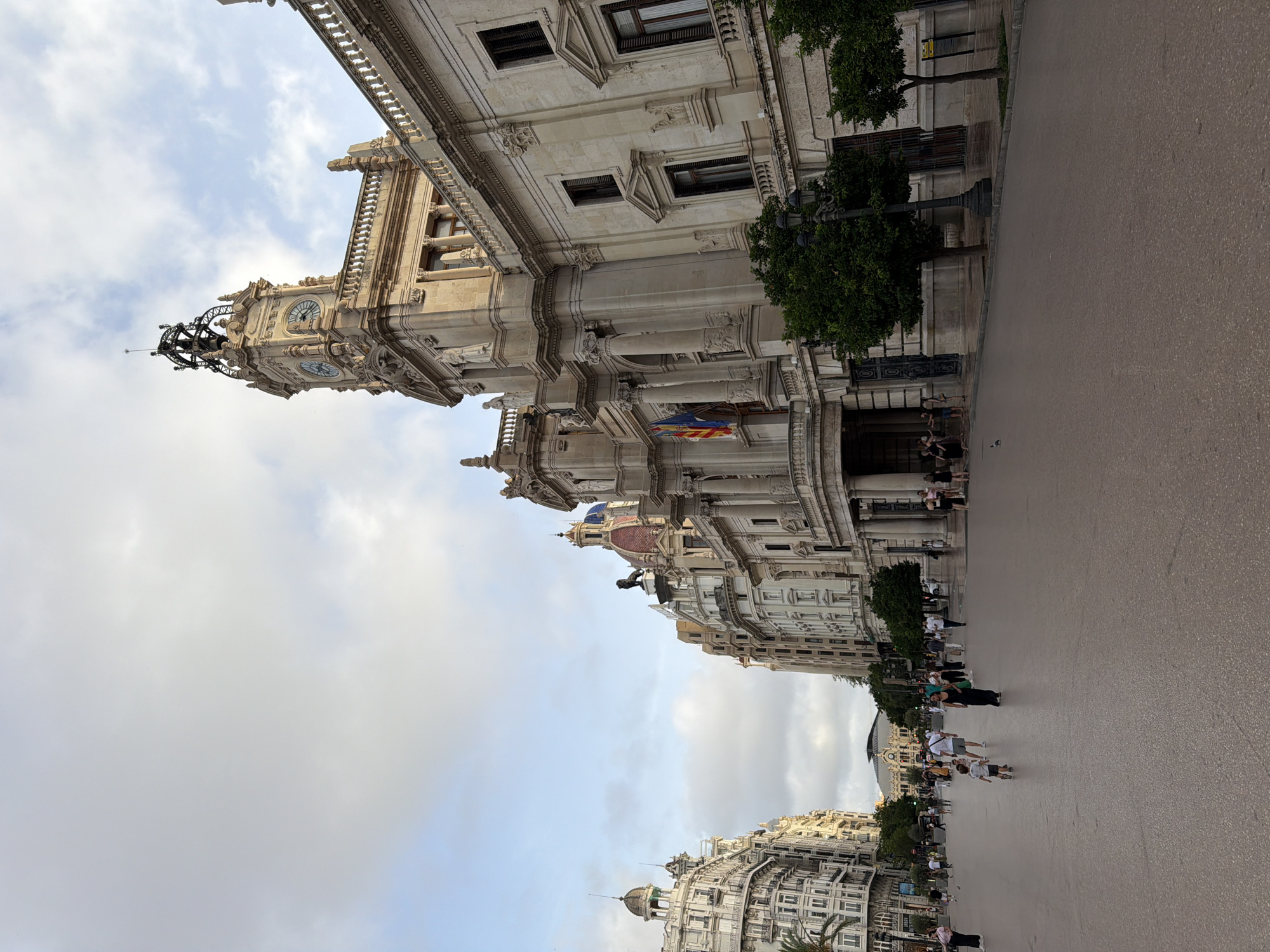An outdoor, eye-level, full shot captures the grandeur of a bustling city square in Valencia, Spain. The foreground is dominated by a wide, grey paved plaza, reflecting the overcast sky above. Several people are scattered across the plaza, some walking, others standing. A young child runs towards the right side of the frame, while a woman in a dark dress walks towards the left.
The mid-ground features a row of imposing, ornate buildings with classical architectural elements, including columns, balconies, and decorative facades. The building on the right is particularly striking, with a prominent clock tower topped by intricate metalwork. Flags, including the Spanish flag, are visible on the facade of one building. Small trees line the sidewalk in front of these buildings, adding a touch of greenery.
In the background, more buildings stretch into the distance, showcasing a consistent architectural style with varied rooflines and window placements. The sky is a pale grey with soft, scattered clouds. The overall impression is one of a vibrant, historic urban center during daylight hours.