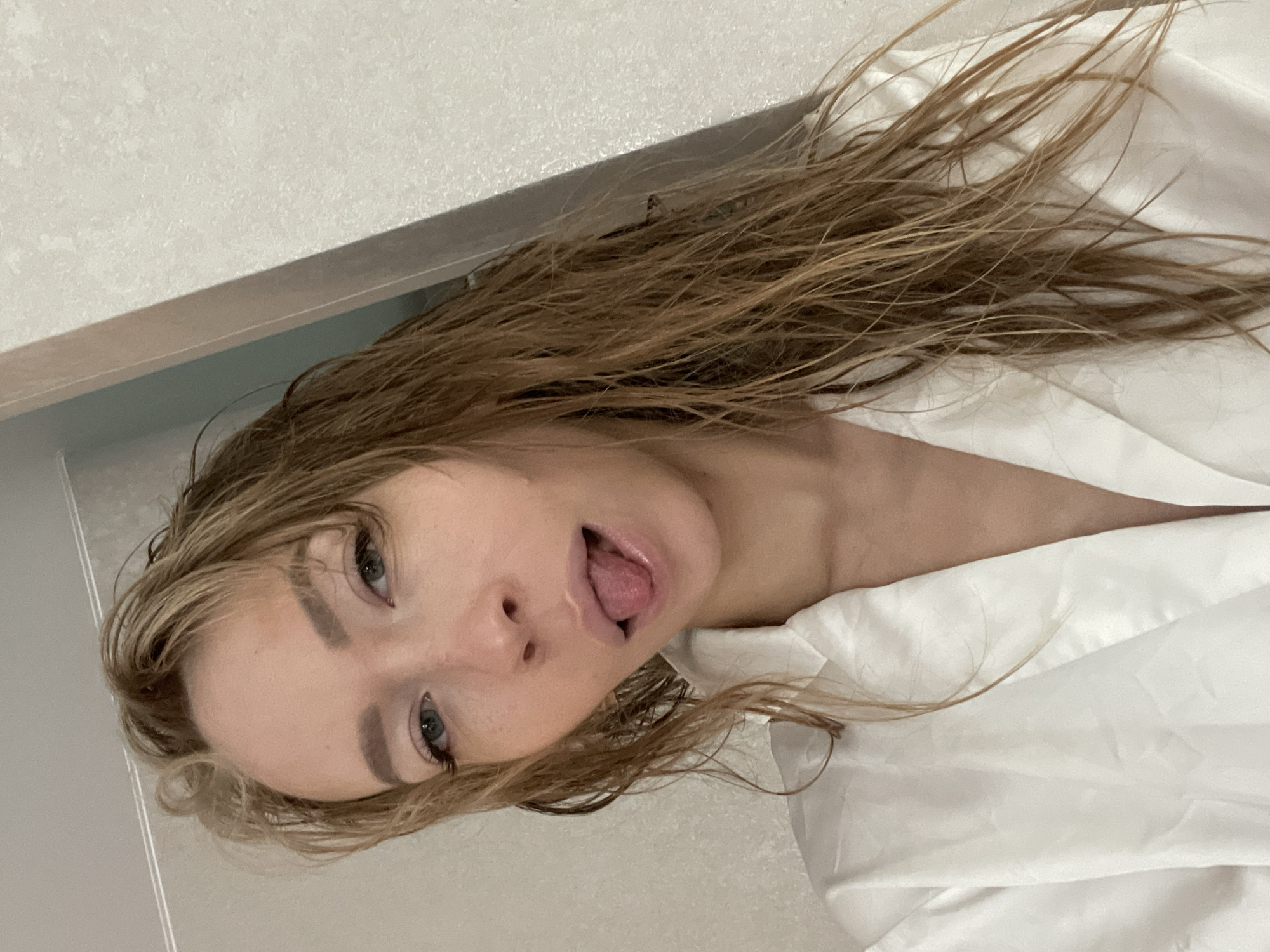 A young woman with wet, blonde hair is sticking her tongue out at the camera. She is wearing a white robe. The background appears to be a bathroom with light-colored walls. There is no discernible location information related to the city of Perm, Russia within the image itself.