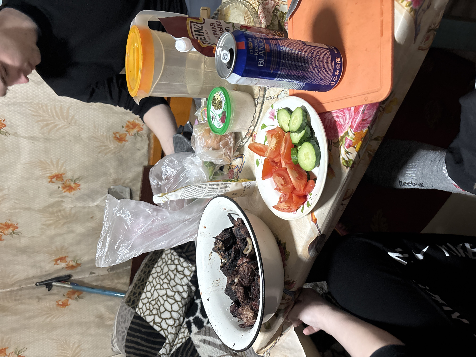 The scene depicts a casual meal setting in what appears to be a private residence.
**Location:** Likely in Chaikovsky, Russia, based on the presence of Cyrillic script on some of the product labels.
**Subjects:** At least two people are partially visible, one wearing a black top and another with grey socks and potentially dark trousers.
**Objects:**
* **Food:** A bowl of what looks like grilled meat, a plate of sliced tomatoes and cucumbers.
* **Drinks:** A can of Kronenbourg Blanc beer.
* **Condiments:** A bottle of Heinz ketchup, a jar of a green condiment.
* **Other items:** A plastic pitcher with an orange top, plastic bags, a cutting board, a patterned tablecloth, a couch/chair with a black and white animal print cover, and an extendable pole leaning against the wall.
**Actions/Interactions:** The people are likely engaged in eating or preparing to eat. The arrangement of the food suggests a shared meal.
**Environment:** The setting appears to be an indoor space, possibly a dining area in a home. The décor suggests a modest and lived-in environment.