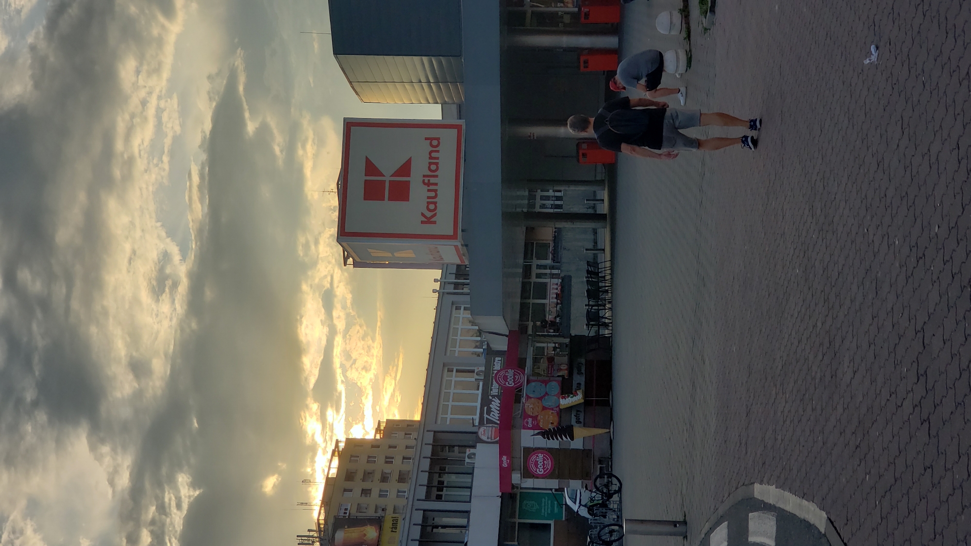 The image depicts a street scene in Prešov, Slovakia, likely near a commercial area, at dusk. The sky is filled with dramatic clouds, illuminated by the setting sun, which casts a warm glow. Dominating the scene is a large "Kaufland" supermarket, identifiable by its prominent red and white signage on the cube-shaped structure. In the foreground, a man wearing a black t-shirt, grey shorts, and a backpack walks away from the camera. To his right, another person, wearing a grey t-shirt and dark shorts, is seated. In the middle ground, a small kiosk with colorful signage, possibly selling ice cream or snacks, is visible. Bicycles are parked to the left of the kiosk. The ground is paved with interlocking bricks, and there are several red trash receptacles scattered throughout the area. Buildings, including a multi-story residential or office building, form the backdrop. The overall impression is of an urban environment winding down for the day under a vibrant sky.
