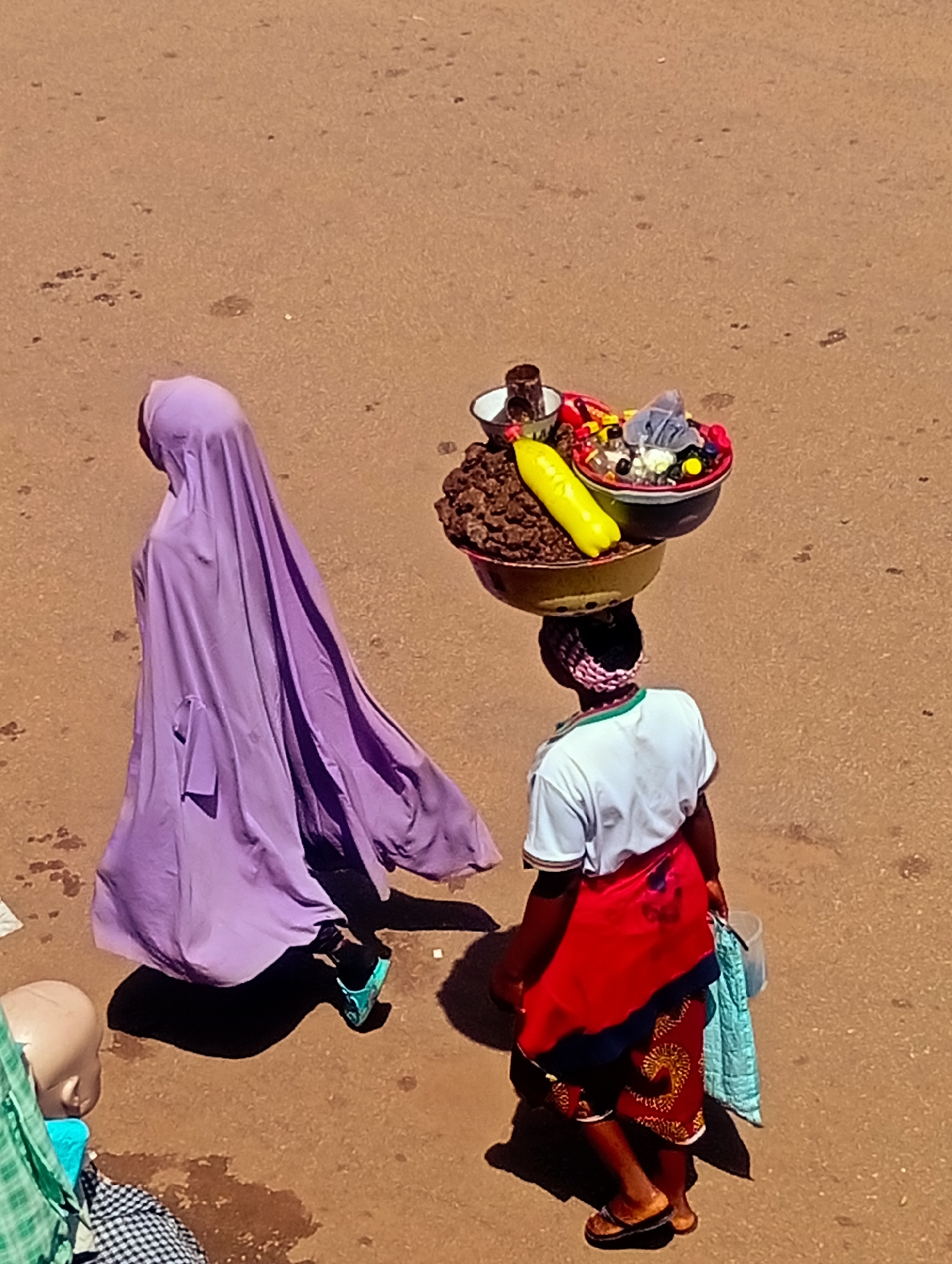 In Bukuru, Nigeria, on a reddish-brown dirt road, two individuals are visible. The person on the left, viewed from behind and in profile, is wearing a long, flowing lavender garment that covers their head and body, with a turquoise shoe visible on their left foot. They are walking away from the viewer. To the right, another person, also viewed from behind and in profile, is carrying a large, tiered basket of goods on their head. The basket contains what appear to be various items, including a long yellow object resembling a banana, small bottles, and dark, granular material. This person is wearing a white t-shirt with a green and white striped collar, a red wrap around their waist, and dark shorts. They are also wearing a patterned headwrap and brown sandals. A light blue cloth is held in their right hand. The scene suggests a marketplace or a street in a populated area, given the presence of the individuals and the items being carried for sale or transport. The ground is dusty with scattered small debris.