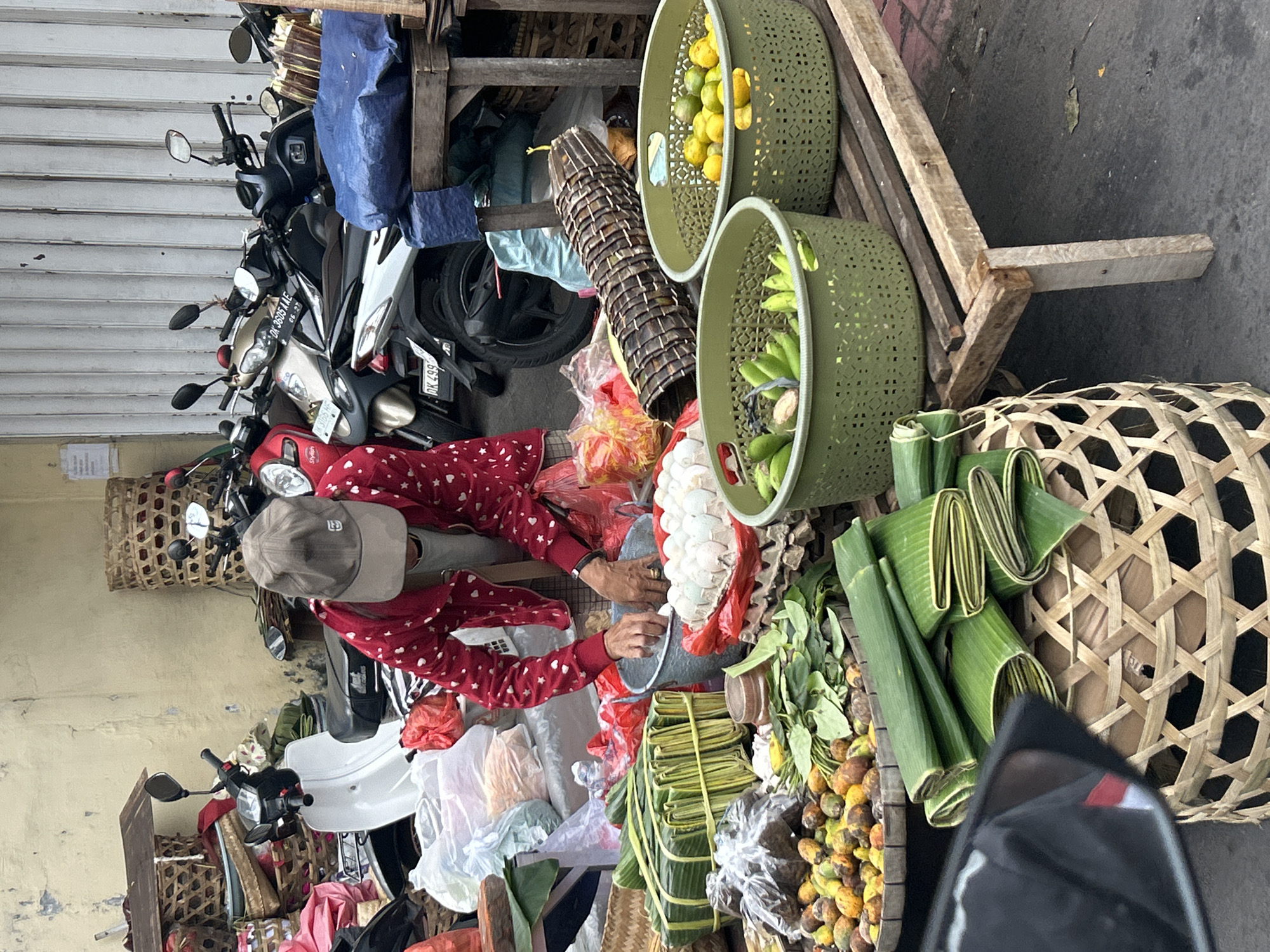 The image depicts an outdoor market scene, likely in Denpasar, Indonesia, given the context of the provided location hint. In the foreground, a person wearing a red jacket with white heart patterns and a grey baseball cap is actively engaged in sorting through items in a grey bucket. This individual's hands are visible as they appear to be handling eggs. Surrounding the person are various market goods, including bundles of green banana leaves, baskets filled with fruits and vegetables such as what appear to be betel nuts, bananas, and citrus fruits. A large woven bamboo tube lies horizontally in the mid-ground. In the background, several motorcycles are parked, suggesting a busy, urban market environment. The ground appears to be asphalt, and a yellow-painted wall forms the backdrop. The overall impression is a vibrant, bustling marketplace.