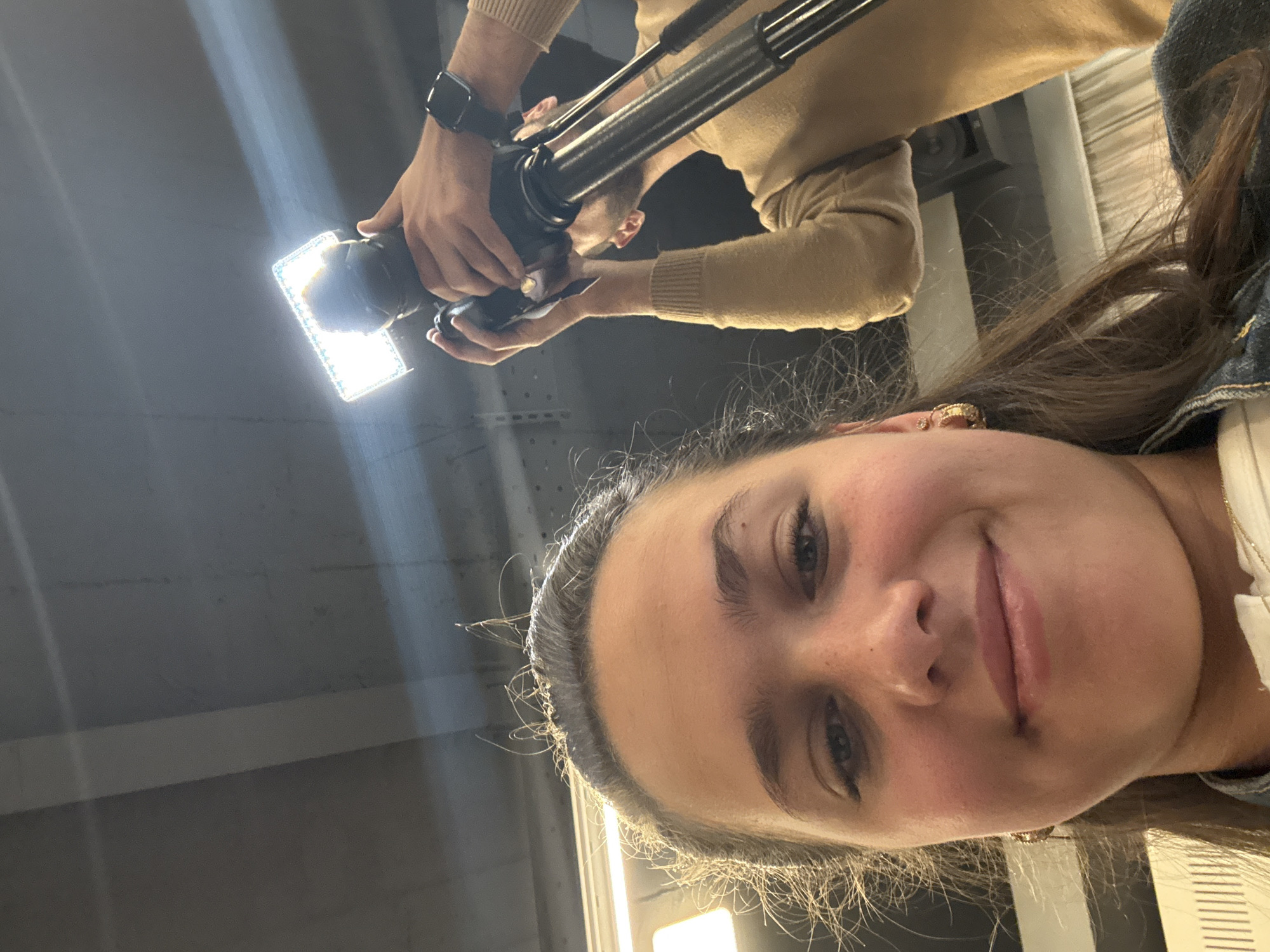 A young woman is taking a selfie in what appears to be a building in the city of Yerevan, Armenia. Above her, a person wearing a light-beige-colored sweater and a smartwatch is holding a camera with a bright light attached to a tripod, as if filming or photographing her. The background appears to be a generic interior setting.