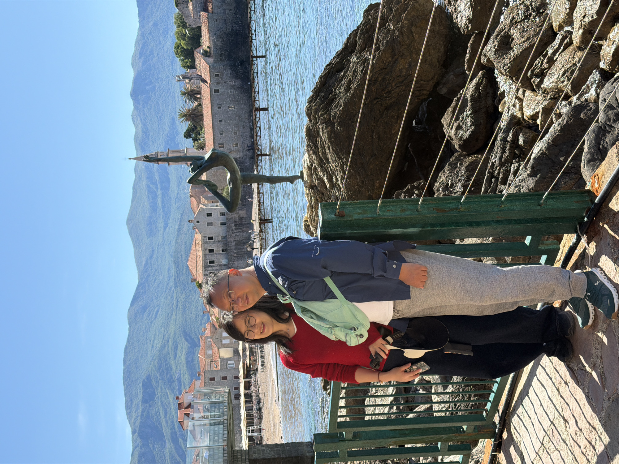 A couple poses for a photo in Budva, Montenegro. The man, wearing a blue jacket and grey sweatpants, stands beside the woman, who is in a red sweater and dark pants. A light green bag is slung across their bodies. They are standing on a stone walkway next to a green metal bench. In the background is the Old Town of Budva, with its buildings, the sea, and mountains. A statue of a dancer is also visible. Some ropes run down the right of the frame.