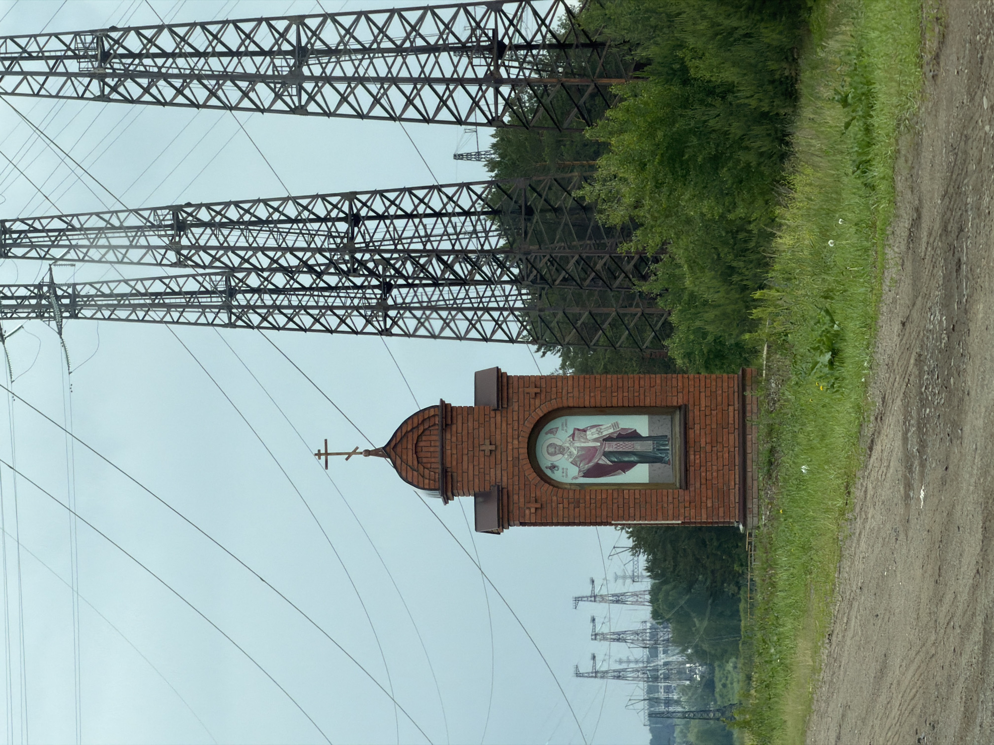 This image, reportedly located in Noviy, Russia, presents a roadside scene featuring a small brick shrine against a backdrop of extensive electrical infrastructure and greenery under an overcast sky.
In the foreground, a light brown dirt road with visible tire tracks occupies the lower left portion of the frame, extending towards the center. To the right of the road and surrounding the base of the shrine is an area of vibrant green grass and various weeds, including some with white dandelion seed heads.
The central subject is a freestanding, reddish-brown brick shrine or chapel, positioned on the left side of the frame. It features a rounded, arch-like roof section topped with a brown Orthodox cross. Below this, a small, darker brown metal roof overhang is present. Within an arched recess on the shrine's front facade, a colorful icon is displayed. The icon depicts a male saint with a halo, dressed in red and gold liturgical vestments, holding a book in his left hand and making a sign of blessing with his right hand.
The background is dominated by large-scale electrical transmission infrastructure. Multiple lattice-steel high-voltage power line towers, appearing dark and weathered, stretch across the middle ground and background, particularly prominent on the right side. Numerous power lines are visible, crisscrossing the light gray-blue sky. Behind the shrine and further into the distance on the left, more transmission towers and associated industrial structures are discernable, partially obscured by a line of dark green trees and shrubs that separate the immediate foreground from the distant structures. Another dense line of trees and shrubs borders the right side of the shrine, extending towards the right edge of the image. The overall impression is a blend of natural landscape, religious monument, and industrial utility infrastructure.