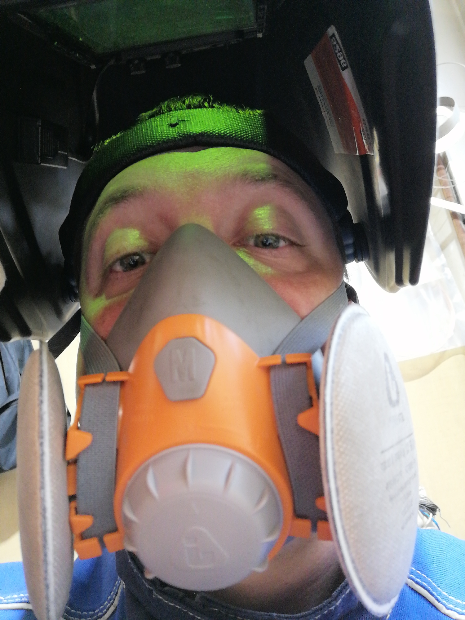 A person is wearing a welding helmet and a respirator mask. The person's face is partially visible, with their eyes looking directly at the camera. The respirator mask is orange and gray. The welding helmet is black. The location is unknown.
