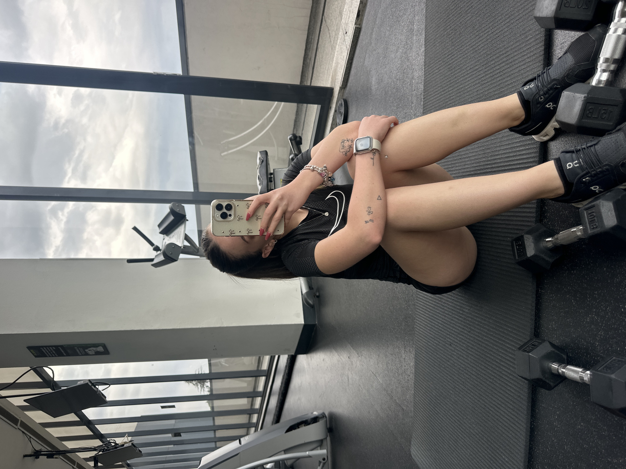 A woman is sitting on a black mat at a gym in Huixquilucan de Degollado, Mexico. She is holding a cell phone in her left hand and taking a photo of herself. She is wearing black shoes, a black shirt, and black shorts. There are dumbbells to her right. A window is behind her, reflecting the sky. Gym equipment is visible in the background.
