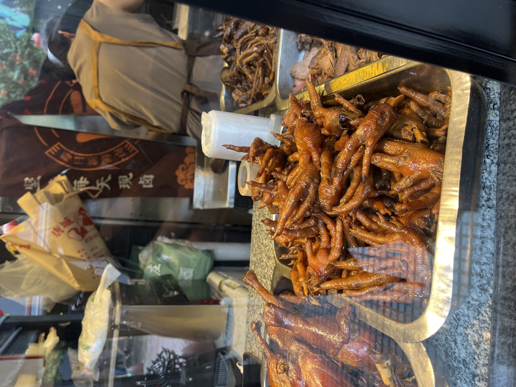 The image displays a vibrant food counter inside an East Asian food establishment, likely located in Buenos Aires, Argentina. The primary subjects are various cooked meats, presented in large metal trays behind a glass display.
In the foreground, a substantial quantity of braised chicken feet fills a rectangular metal tray. They exhibit a rich, glossy reddish-brown hue, characteristic of being cooked in a savory soy-based sauce. Below this, another tray contains whole cooked ducks or other poultry, similarly dark brown and appearing to be braised or roasted. Further back, additional trays hold other cuts of dark, cooked meat, possibly offal or different types of braised dishes.
A roll of clear plastic bags, presumably for packaging purchases, is visible near the chicken feet. The background shows a bustling scene through the glass. A person wearing a light grey top with gold trim is partially visible on the upper left. Below them, a dark brown banner or fabric sign displays prominent Chinese characters: "滷味" (lǔ wèi), meaning "braised/stewed dishes," and "熟食" (shú shí), meaning "cooked food." Part of another character, possibly "臺灣" (Taiwan), is also discernible. To the left, several plastic and paper bags are visible, one beige paper bag clearly showing "唐城" (Tángchéng), which translates to "Tang City" or "Chinatown." The counter surface beneath the trays is a speckled grey, resembling granite. The scene suggests a busy deli or take-out section offering a variety of traditional cooked dishes.