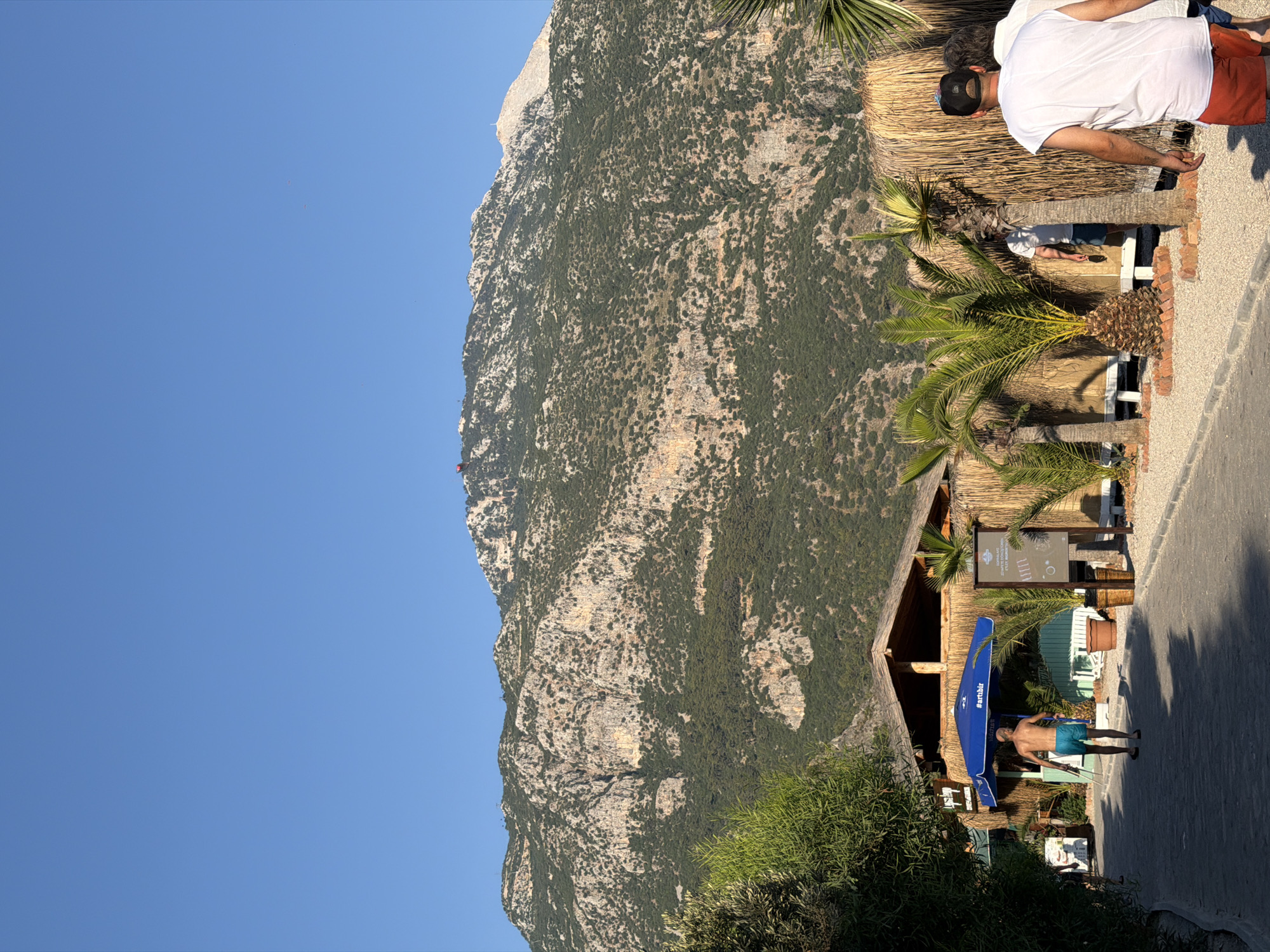 A high-angle, wide shot captures a sunlit, dusty path leading towards a rustic beachfront establishment in Fethiye, Turkey, with a massive, verdant mountain dominating the background under a clear blue sky. In the foreground, the path is lined with palm trees and a thatched-roof building adorned with a large blue umbrella bearing the "#artibir" hashtag. Several individuals are visible: a shirtless man in blue swim trunks walks away from the camera on the path, appearing to head towards the building; further back, a person in a white shirt and red shorts walks towards the camera, their back to the viewer; another person in a white shirt is partially visible behind a palm tree; and a person is seen walking away from the camera on the far left edge of the frame. The establishment itself features a straw-covered wall, potted plants, and outdoor seating areas. The mountain in the background is rugged and covered in dense green foliage, with signs of rock erosion and clear geological strata. A small red structure is visible near the peak of the mountain, and what appears to be a weather station or a marker stands on another part of the summit. The lighting suggests it is either early morning or late afternoon, casting long shadows across the scene.