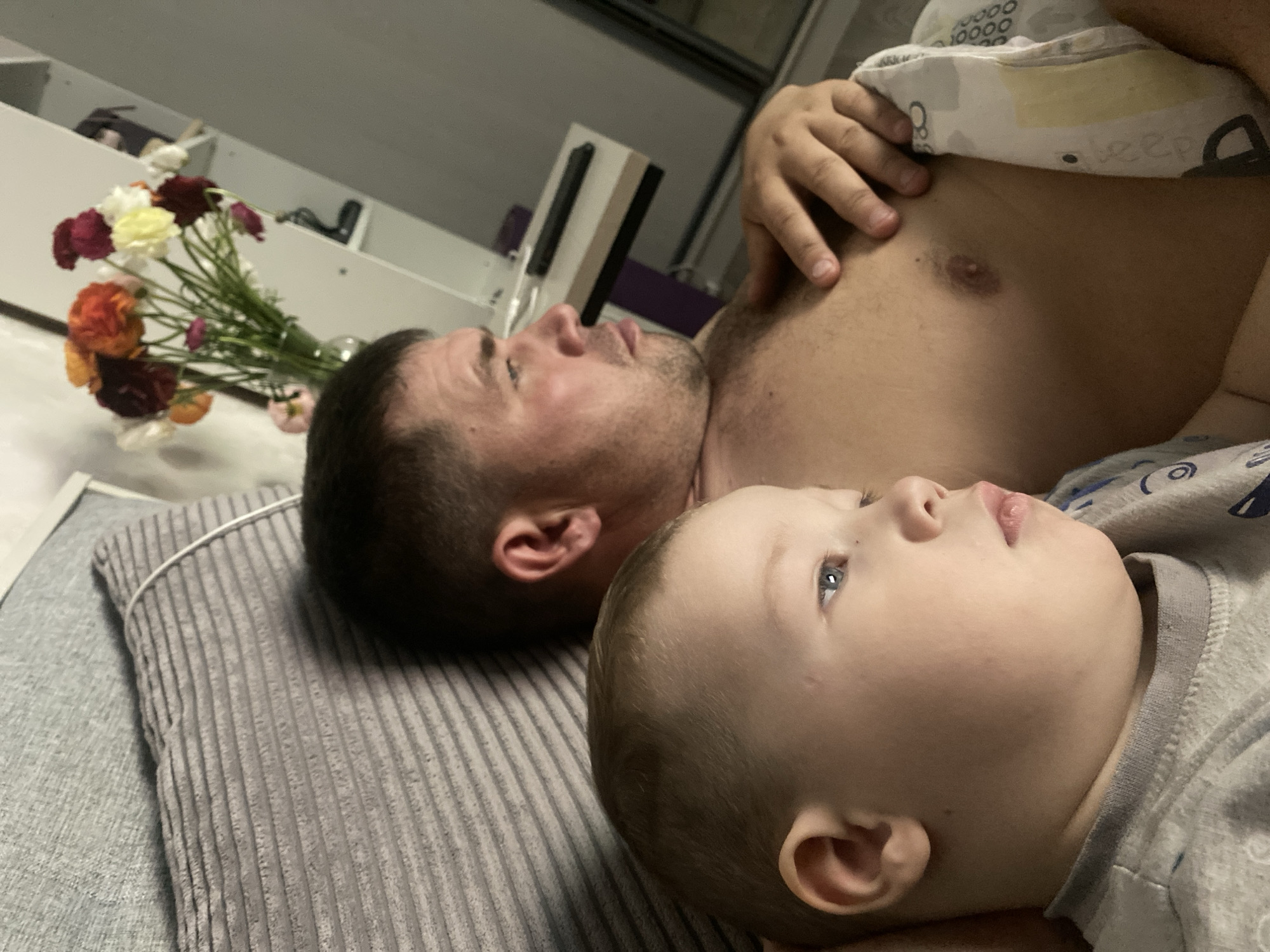 An intimate, upward-angled shot captures an adult man and a young boy lying side-by-side, likely in a bedroom or living space, in Sochi, Russia. Both individuals are positioned on their backs, with their heads turned to their left, gazing upwards in the same direction, suggesting a shared point of interest or a moment of quiet contemplation.
The adult man, shirtless and with stubble, is partially covered by a light-colored blanket adorned with a repeating pattern of circles and the word "sleep." His right hand rests near his shoulder. The young boy, with light hair and clear blue eyes, lies directly beside him, wearing a grey patterned top. Their expressions are calm and relaxed, conveying a sense of peace and closeness.
To the left of the man's head, a vibrant bouquet of flowers in various hues including red, orange, yellow, white, pink, and purple, sits in a clear vase, adding a touch of domesticity to the scene. They are positioned on a light surface, possibly a bedside table or shelf. The man rests his head on a grey, ribbed pillow, which in turn lies on a lighter grey flat surface. The background is an indistinct indoor setting with some shelves or furniture visible. The lighting is soft and even, creating a gentle atmosphere for this shared moment between father and son.