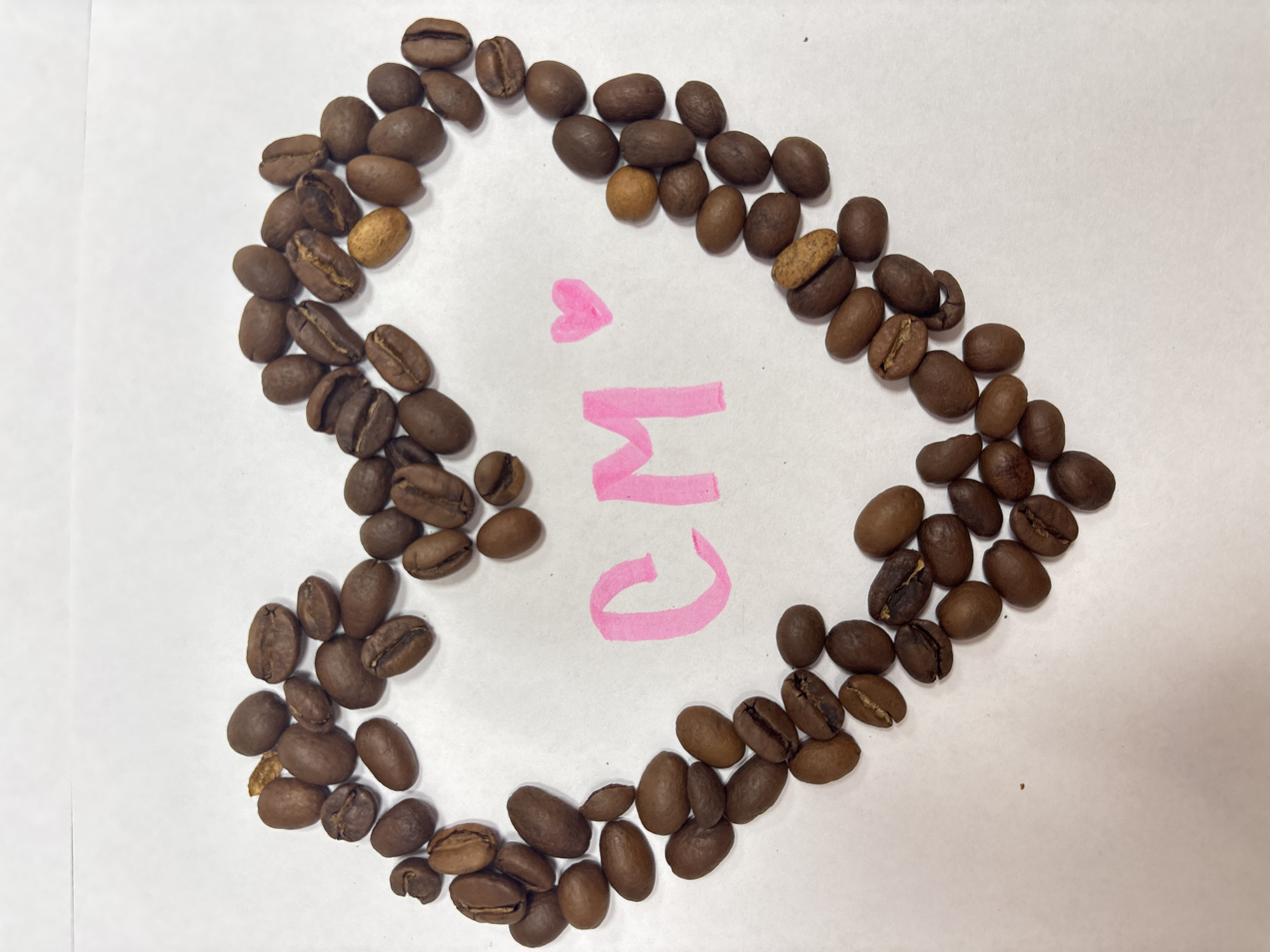 A top-down shot shows a collection of roasted coffee beans arranged in a heart shape on a white surface. Inside the heart, the pink letters "CM" are written, accompanied by a small pink heart. The beans are various shades of brown, with a few lighter-colored beans scattered throughout. The setting is a simple, undetailed white background. The image does not contain any information that suggests that it was taken in the city of Chaikovsky, Russia.