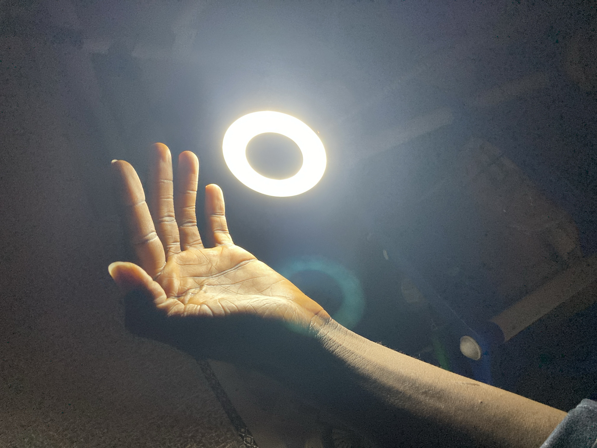 The image shows a close-up of a person's outstretched hand and forearm against a dark background. A bright, circular ring light is positioned above and slightly behind the hand, casting a warm glow on the skin and creating a strong backlight effect. The light illuminates the palm and fingers, highlighting the texture of the skin and casting shadows. A subtle blue lens flare is visible to the right of the hand. The background is dark and indistinct, with some areas of lighter gray suggesting a textured surface, possibly a wall or fabric. The overall mood is one of focused attention, with the light drawing the viewer's eye to the hand. The context of Jos, Nigeria, suggests an indoor setting, likely at night or in a dimly lit space, given the prominent artificial light source. There is no visible text in the image.