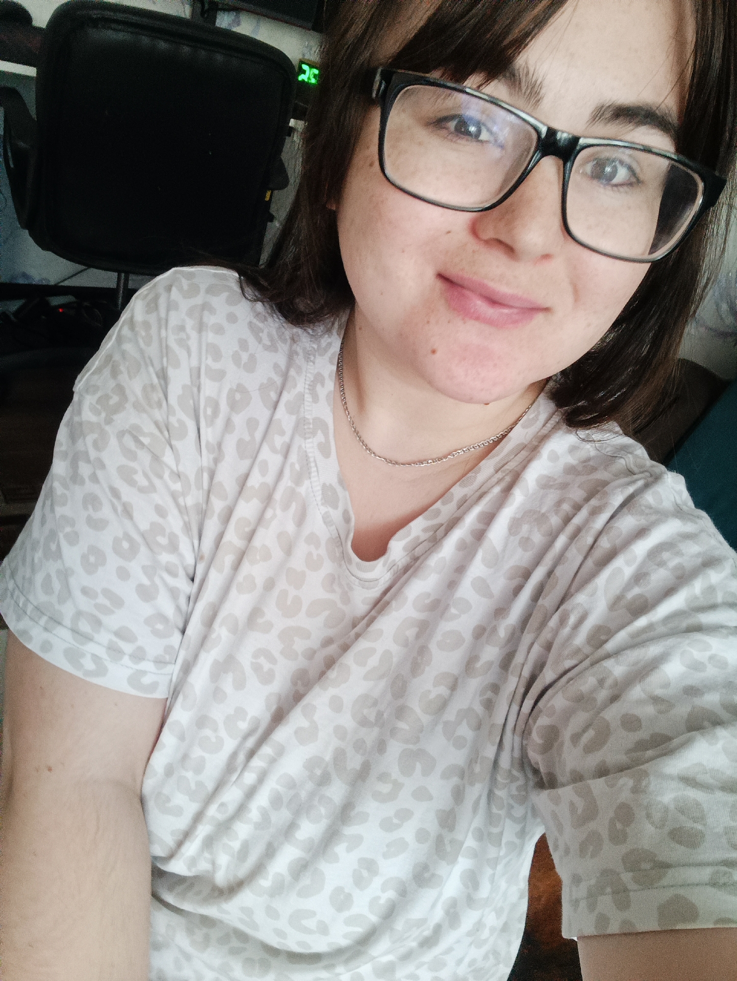 This close-up, chest-up selfie features a young woman with dark hair and dark, thick eyebrows, looking directly at the camera with a subtle, closed-mouth smile. She has fair skin with visible freckles across her cheeks and chin. She wears prominent black-framed glasses, through which her eyes are visible, reflecting some light on the left lens. Her attire includes a casual, light-colored t-shirt adorned with a beige or light brown leopard print pattern. A delicate silver-toned chain necklace rests around her neck.
The background is softly blurred but reveals elements of an indoor setting. To the woman's left, a segment of a black office chair is visible. Further back, a device with glowing green digits displaying "25" can be seen. Other obscured shapes suggest a typical room environment, possibly with electronics. The lighting is soft and even. The overall impression is one of calm and casualness. The image is provided with the location context of Noviy, Russia, although no specific visual cues within the photo confirm this.