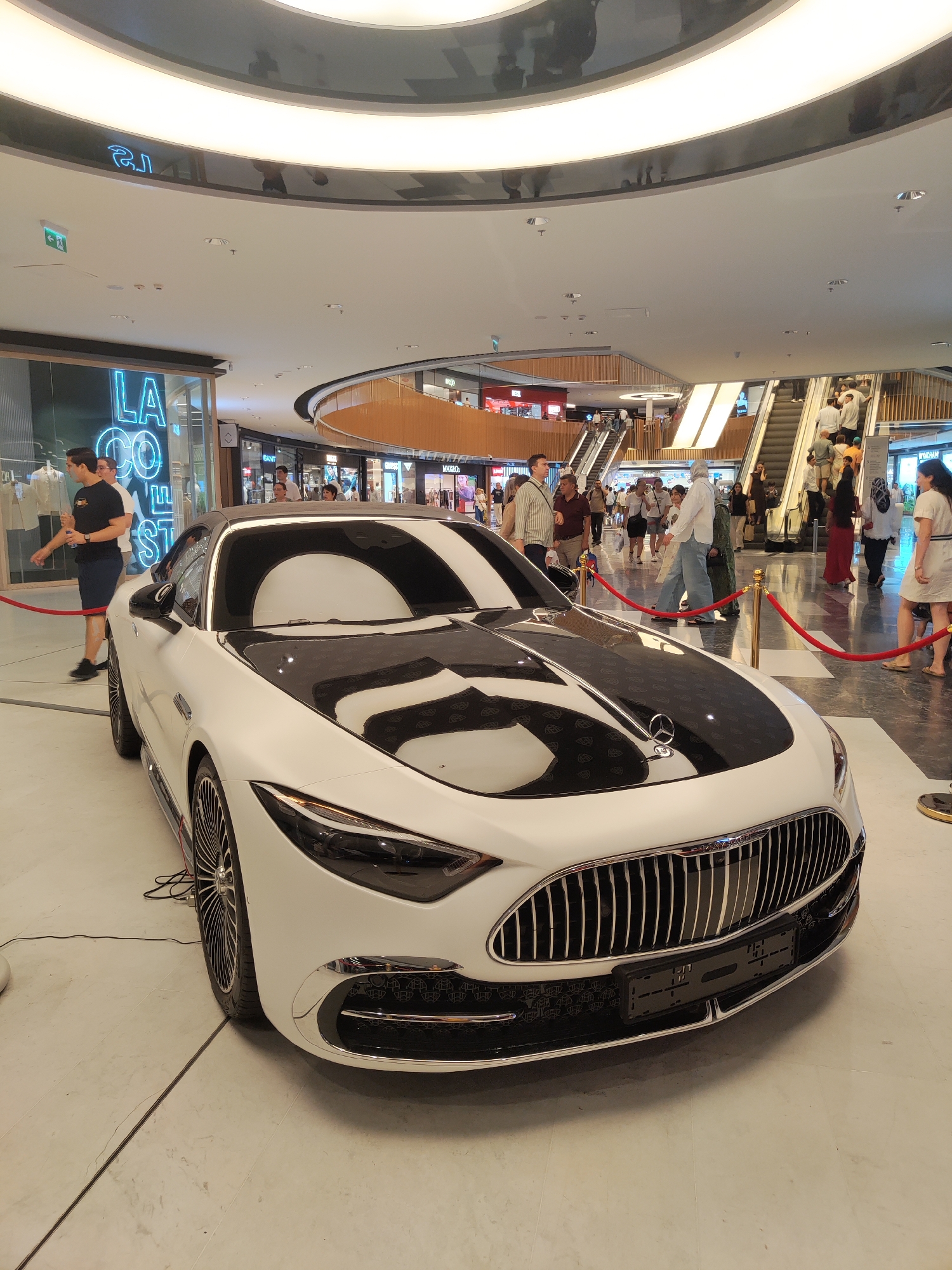 A white Mercedes-Benz Maybach is on display in a modern shopping mall. The car has a black hood with a patterned design and chrome accents. It is cordoned off with red velvet ropes. In the background, there are escalators with people walking up and down, as well as various shop fronts, including one with a blue neon sign that says "LA CO". The mall has a high, curved ceiling with reflective panels. The flooring is light-colored tile. The scene appears to be in Tashkent, Uzbekistan.