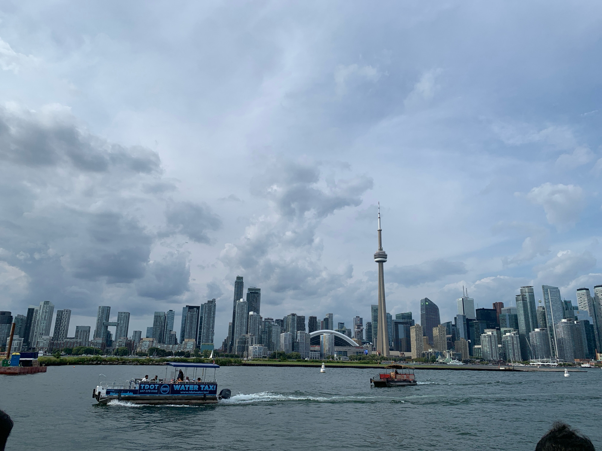 This is an aerial view of the Toronto skyline and waterfront on an overcast day. In the foreground, two boats are traveling on the water. The boat on the left is a white water taxi with "TDOT WATER TAXI" written on its side, carrying several passengers. The boat on the right is smaller and appears to be a workboat. The skyline of Toronto is visible in the background, featuring numerous skyscrapers of varying heights and architectural styles. Prominently displayed is the CN Tower, a tall, slender structure. Beyond the buildings, the sky is filled with clouds, with patches of lighter blue showing through. The water appears choppy, indicating movement from the boats and wind.
