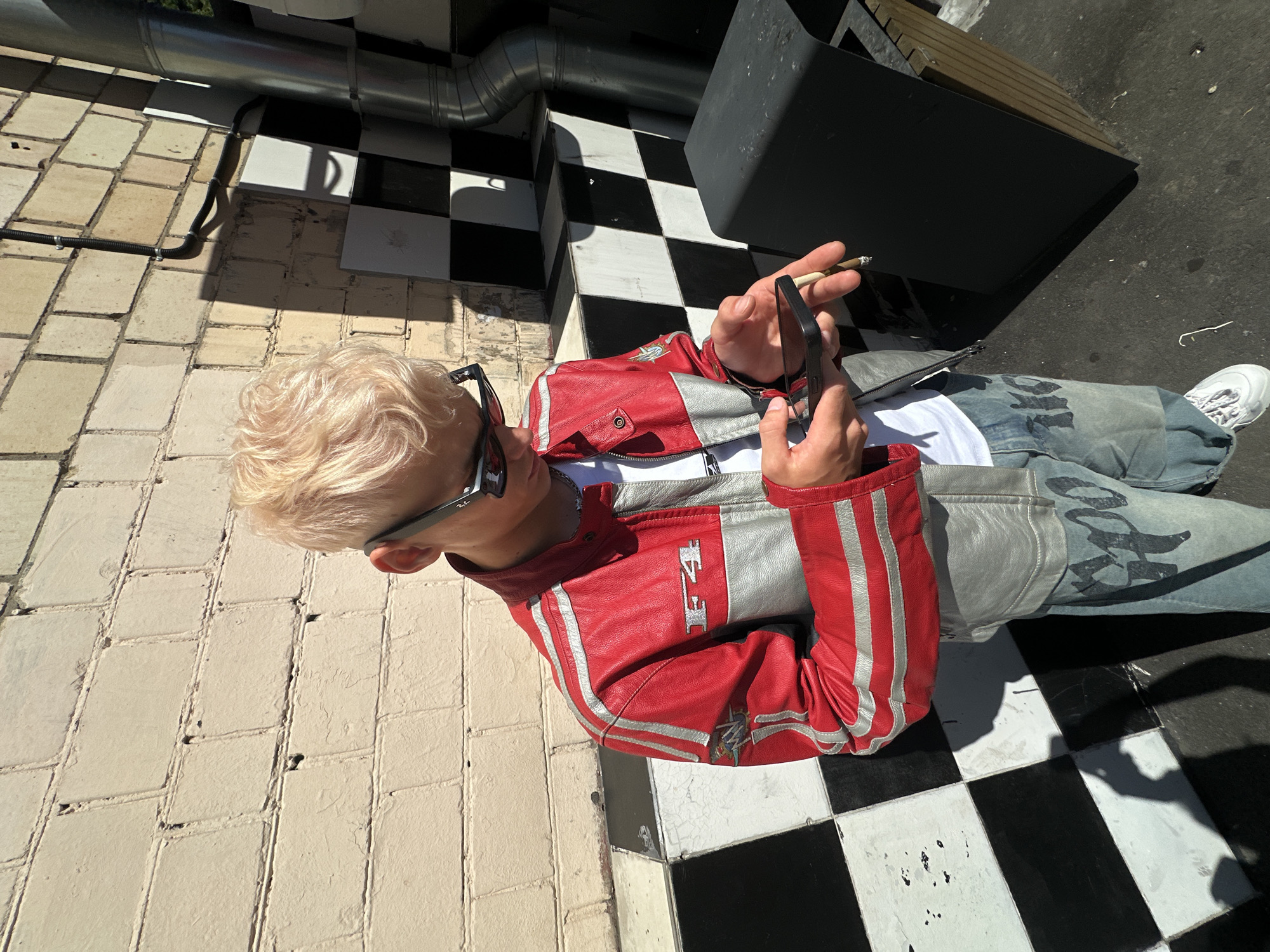 A young person with bleached blonde hair and sunglasses, wearing a red and silver racing jacket, a white t-shirt, and light-wash jeans with black graphics, stands outdoors in front of a brick wall with a checkerboard pattern. They are holding a smartphone in their left hand and a cigarette in their right, looking down at the phone. The background includes a metal pipe and a black trash bin. The lighting suggests it is a sunny day. The location is identified as a street in Moscow, Russia.