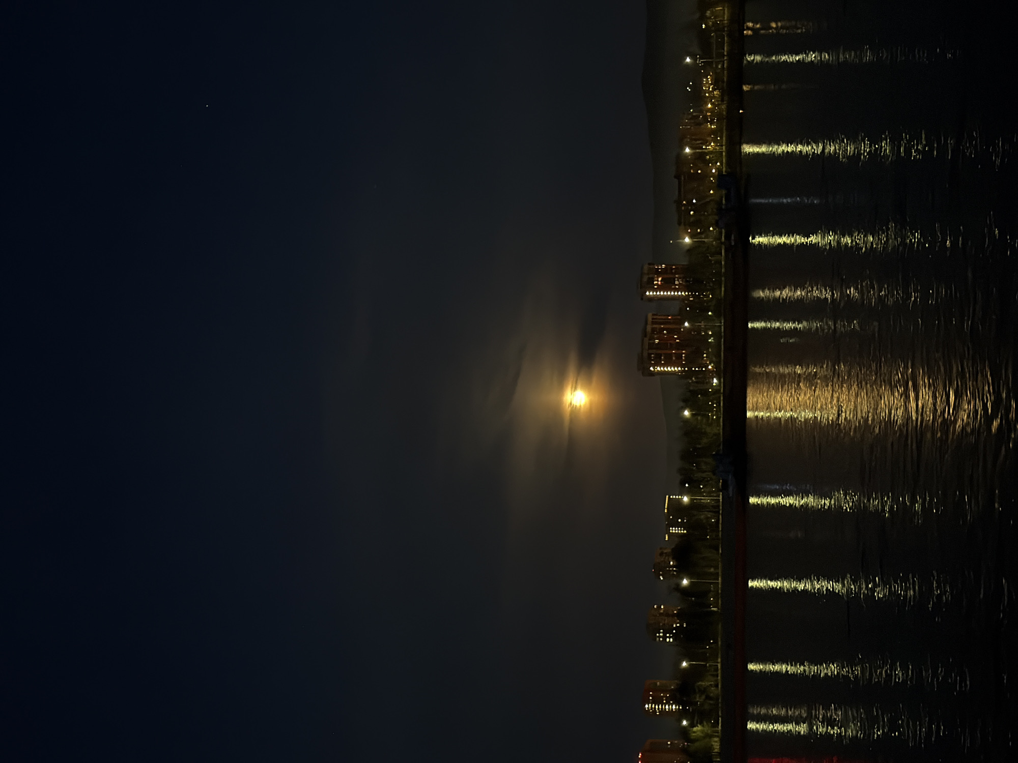 The image presents a serene night scene of a city illuminated by both artificial lights and a prominent full moon, viewed across a body of water. The composition is horizontally split between the dark sky and land on the left, and the reflective water on the right.
In the upper-left portion of the dark, deep blue night sky, a large, bright full moon dominates, casting a strong yellow-orange glow. It appears partially veiled by thin clouds, which diffuse its light and create soft, radial streaks. A tiny, faint white speck, possibly a star or distant planet, is barely visible further into the dark sky, away from the moon's brilliance.
Along the left edge of the image, the dark silhouettes of a city or landmass are visible. Several multi-story buildings, likely residential or commercial, stand near the water's edge, their numerous windows glowing warmly with yellow light. Scattered streetlights or promenade lights add more points of illumination. In the far background, the indistinct outline of dark hills or mountains rises against the night sky, suggesting a mountainous or hilly setting for the city.
The right side of the image is occupied by the dark, placid water, which acts as a natural mirror. It beautifully reflects the vibrant yellow and orange lights from the city buildings, streetlights, and the moon above. These reflections appear as elongated, shimmering streaks across the water's surface, indicating gentle ripples or a prolonged photographic exposure capturing the movement.
The time of day is clearly night, evidenced by the darkness of the sky and the widespread artificial illumination. The overall atmosphere is tranquil and captivating, showcasing the urban landscape under celestial light. No specific activity or visible text can be identified in the image. Given the context, this could be a view of Krasnoyarsk, Russia.