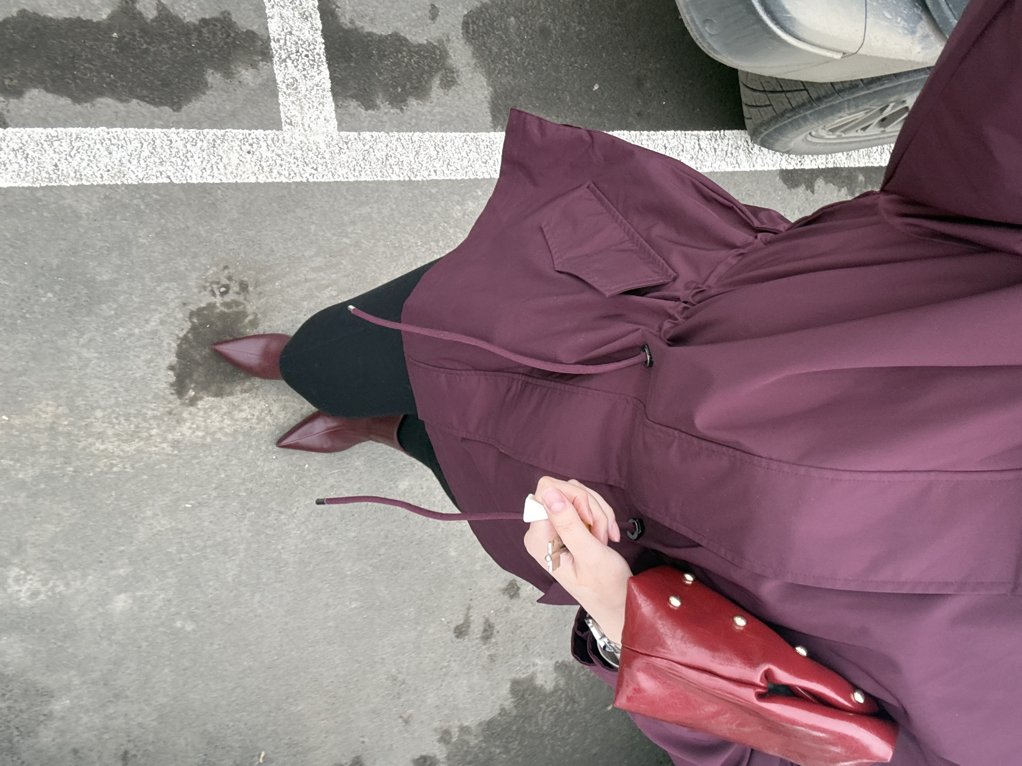 A high-angle, close-up shot shows an individual standing on a paved surface, possibly concrete or asphalt, with white parking lines visible. The person is wearing black leggings, burgundy pointed-toe boots, and a long, plum-colored coat with a drawstring. The coat has a large pocket flap on the upper portion and a visible drawstring. A red patent leather handbag with gold studs is slung over the person's shoulder. The person's left hand is visible, holding a key with a white keychain. Part of a silver car, including a tire, is visible in the upper right corner of the image. The location is indeterminate, with no visible landmarks. It is not possible to confirm it is in Izhevsk, Russia.