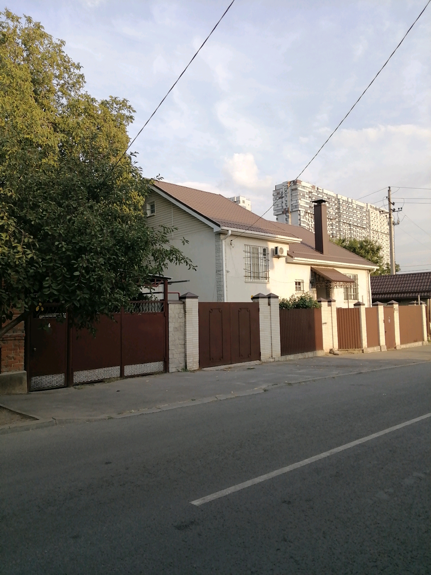 This is a photograph of a street scene in Rostov-on-Don, Russia, with a residential house and a tall apartment building in the background. The foreground shows a grey asphalt road with a white line marking the lane. A sidewalk runs alongside the road, bordered by a fence. The fence is made of alternating brick pillars and dark brown metal panels. A large green tree with dense foliage is on the left side of the image, partially obscuring a brick wall. The house is a single-story structure with a brown roof, white siding, and windows with metal bars. An air conditioning unit is visible on the exterior wall. In the distance, a large, modern apartment building with many windows dominates the skyline. Power lines are visible crisscrossing the sky. The lighting suggests it is late afternoon or early evening, with the sun casting soft shadows. There are no people visible in the image.