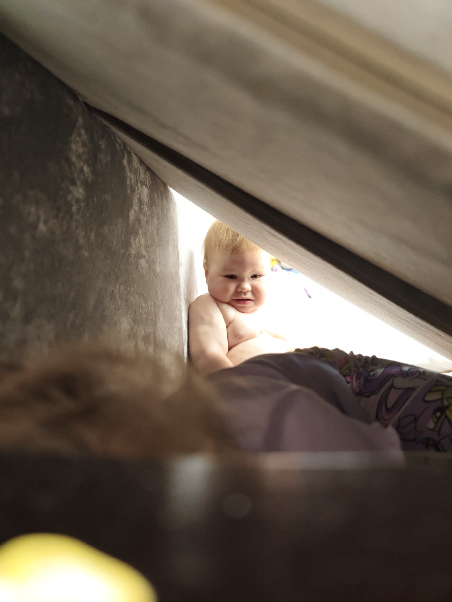 A partially visible Caucasian baby with blond hair is sitting under a structure made from grey fabric in what appears to be a home in Chaikovsky, Russia. The structure is angled towards the baby. The baby has a slightly furrowed brow. A patterned fabric is partially covering the baby's lower half. Some blurred objects are in the foreground.
