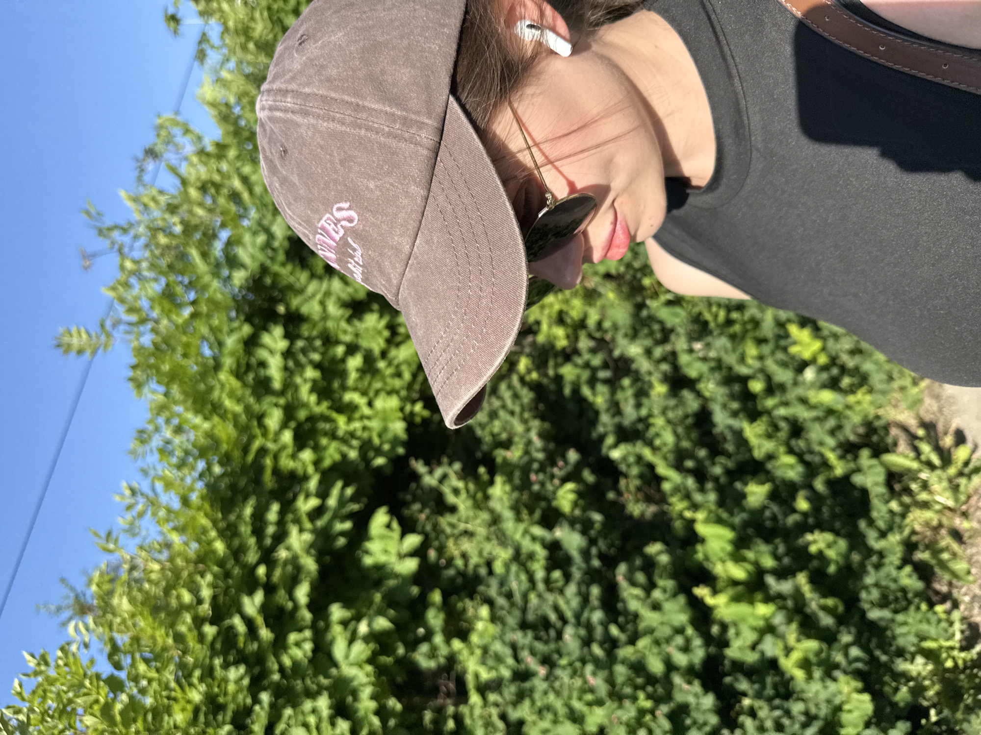 A young woman wearing a brown baseball cap and sunglasses is outdoors, facing to the right, with a dense green hedge behind her and a clear blue sky above. She has a white wireless earbud in her right ear and is wearing a dark, sleeveless top. A brown strap, possibly from a bag, is visible over her shoulder. The lighting suggests it is daytime. There are no visible landmarks that specifically place the scene in Szklarska Poręba, Poland.