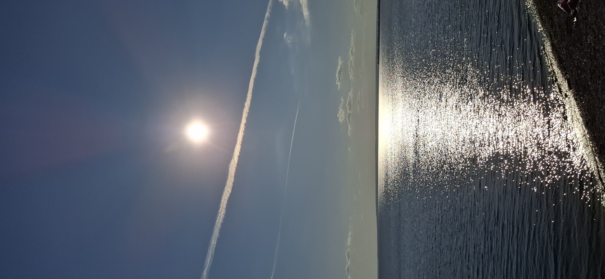 The image depicts a bright, sunny day over a body of water, likely the Kuibyshev Reservoir in Tolyatti, Russia. The sun is high in the clear blue sky, casting a brilliant reflection on the rippling water, creating a shimmering path of light. A faint contrail stretches across the sky from the upper left to the lower right. In the distance, a hazy shoreline is visible, hinting at the vastness of the reservoir. The foreground shows a strip of pebbly ground, suggesting the image was taken from the shore. No people or specific objects are clearly identifiable in the scene, creating a peaceful and expansive natural landscape.