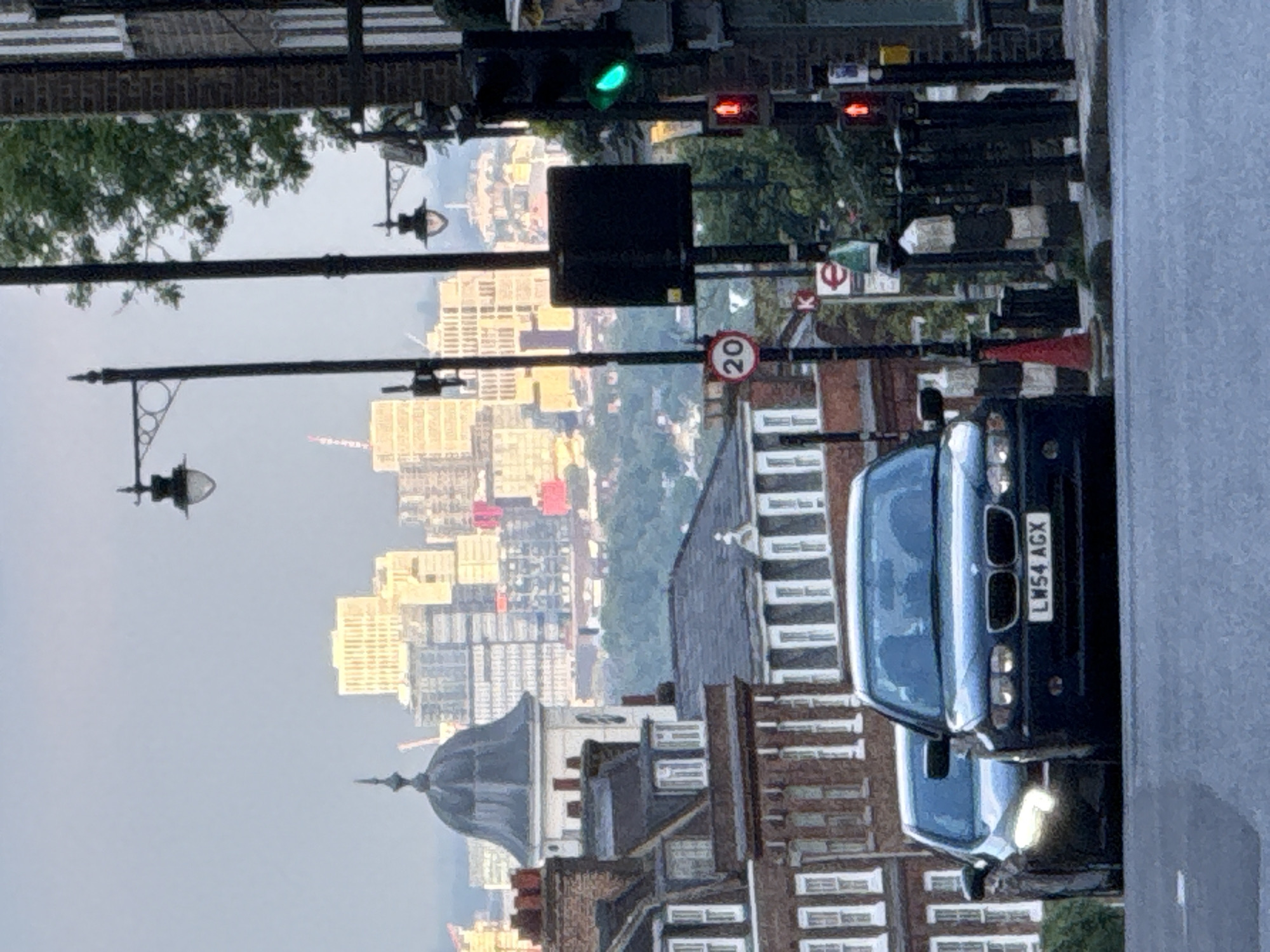 The image depicts a street scene in London, United Kingdom. In the foreground, a black car is parked on the side of the road. The license plate reads "LM54 AGX". Adjacent to the car, there is a 20 mph speed limit sign. A street lamp stands prominently in the middle ground. Beyond the street lamp, there is a view of a city skyline with various buildings and skyscrapers visible. A green traffic light is active above the traffic and street signs. In the far background, there is a glimpse of what appears to be the River Thames. Buildings and residential housing in the area appear well-kept and orderly. The weather seems overcast.