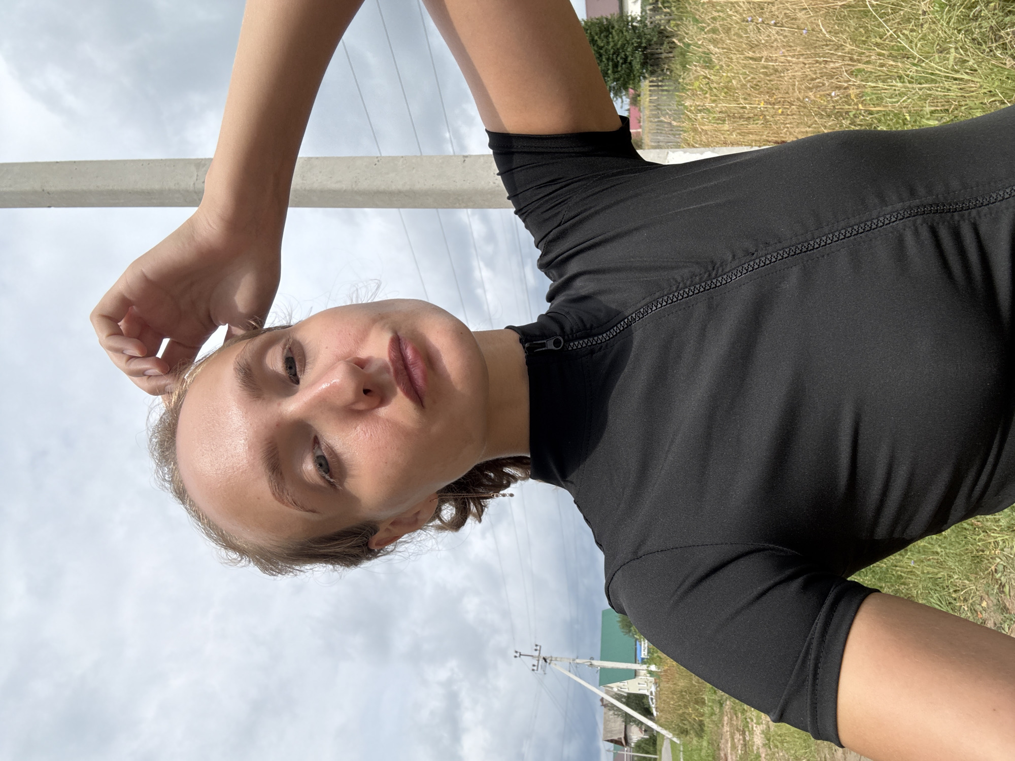 A young woman with blonde hair is taking a selfie outdoors in the village of Podshivalovo, Russia. She is wearing a black, short-sleeved, zippered top. The background shows a cloudy sky, a concrete utility pole, rural buildings, and dry grass. The woman is touching her hair with her right hand, and her left arm is bent at the elbow.