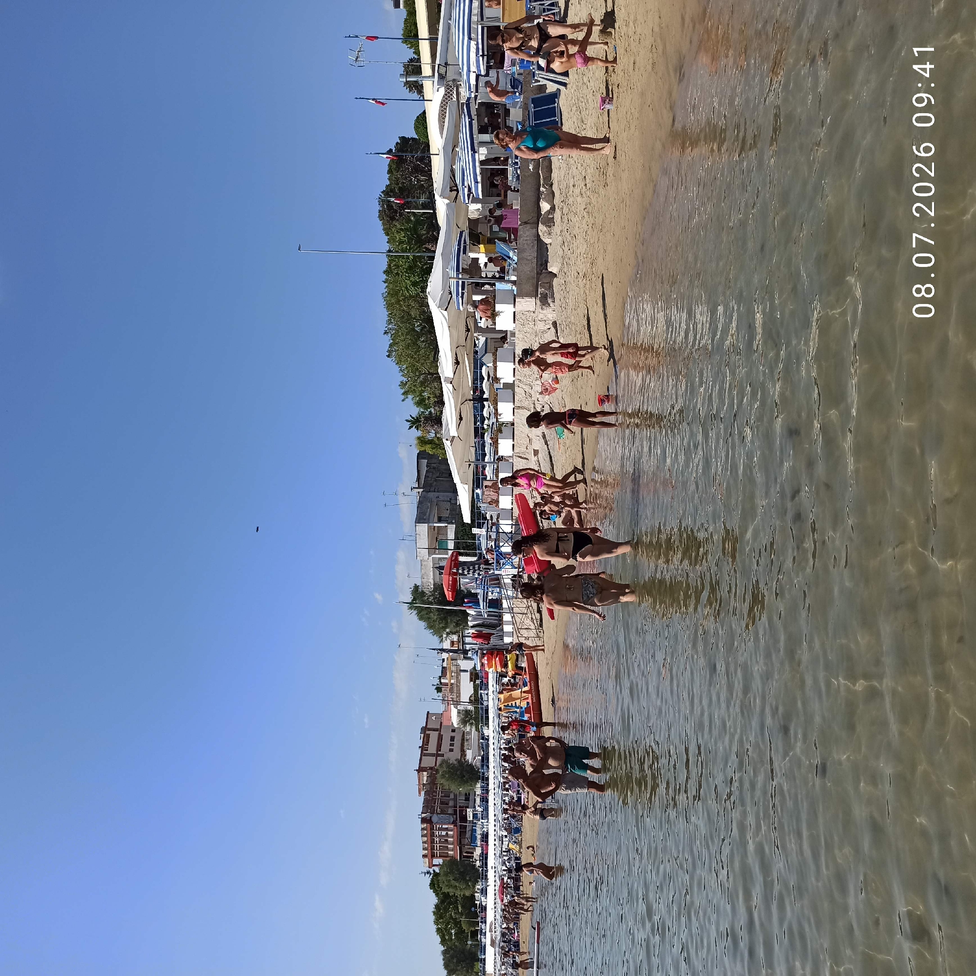 A wide, low-angle shot captures a lively beach scene in Trani, Italy, on a sunny day. The foreground is dominated by shallow, clear turquoise water with sunlight glinting off the sandy bottom. Several people are wading in the water, their lower bodies submerged.
The sandy beach is populated with numerous individuals, many in swimwear, enjoying the warm weather. Beach umbrellas provide shade for some, while others are bathing in the sea or walking along the shore. A prominent red lifeguard boat is positioned on the sand, indicating a supervised beach area.
In the mid-ground, a strip of beach features loungers and more umbrellas, with a few buildings visible in the background, suggesting the presence of hotels or beach facilities. Trees and vegetation line the edge of the developed area.
The sky is a clear, bright blue with a few wispy clouds. A small, dark object, possibly a bird or drone, is visible high in the sky. The date stamp "08.07.2026 09:41" is present in the lower right corner of the image. The overall atmosphere is one of a typical summer day at a popular seaside resort.