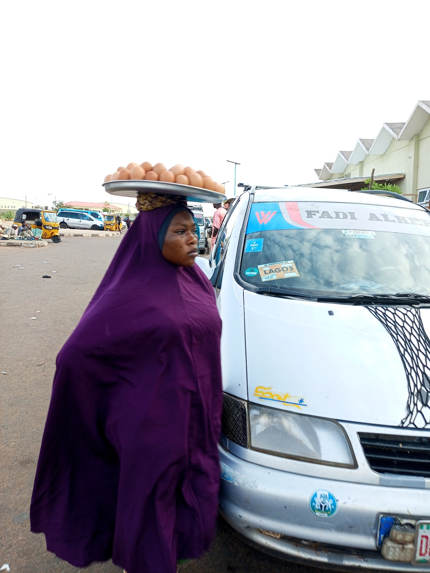 In Gombe, Nigeria, a woman wearing a vibrant purple hijab walks with a large metal tray of eggs balanced on her head. She is positioned in the foreground, walking on a paved street, with the side of a silver minivan prominently featured to her right. The minivan has various stickers on its windshield, including one that reads "FADI ALHERI" and another with "LAGOS." In the background, other vehicles, including yellow tricycles, and buildings are visible, suggesting a busy urban environment. The sky is overcast.