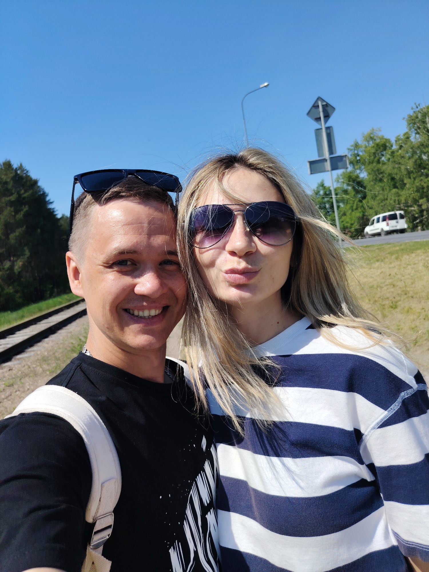A selfie photograph depicts a man and a woman standing outside on a sunny day. The location is Chaikovsky, Russia. The man is positioned on the left of the frame, wearing a black t-shirt with a white graphic design, a white backpack, and sunglasses perched on his head. He is smiling. The woman is on the right, wearing a blue and white striped long-sleeved shirt and dark sunglasses. She is puckering her lips for a kiss. In the background are railroad tracks to the left, a grassy area, a road with a white van, trees, a street lamp, and a road sign. The sky is blue.