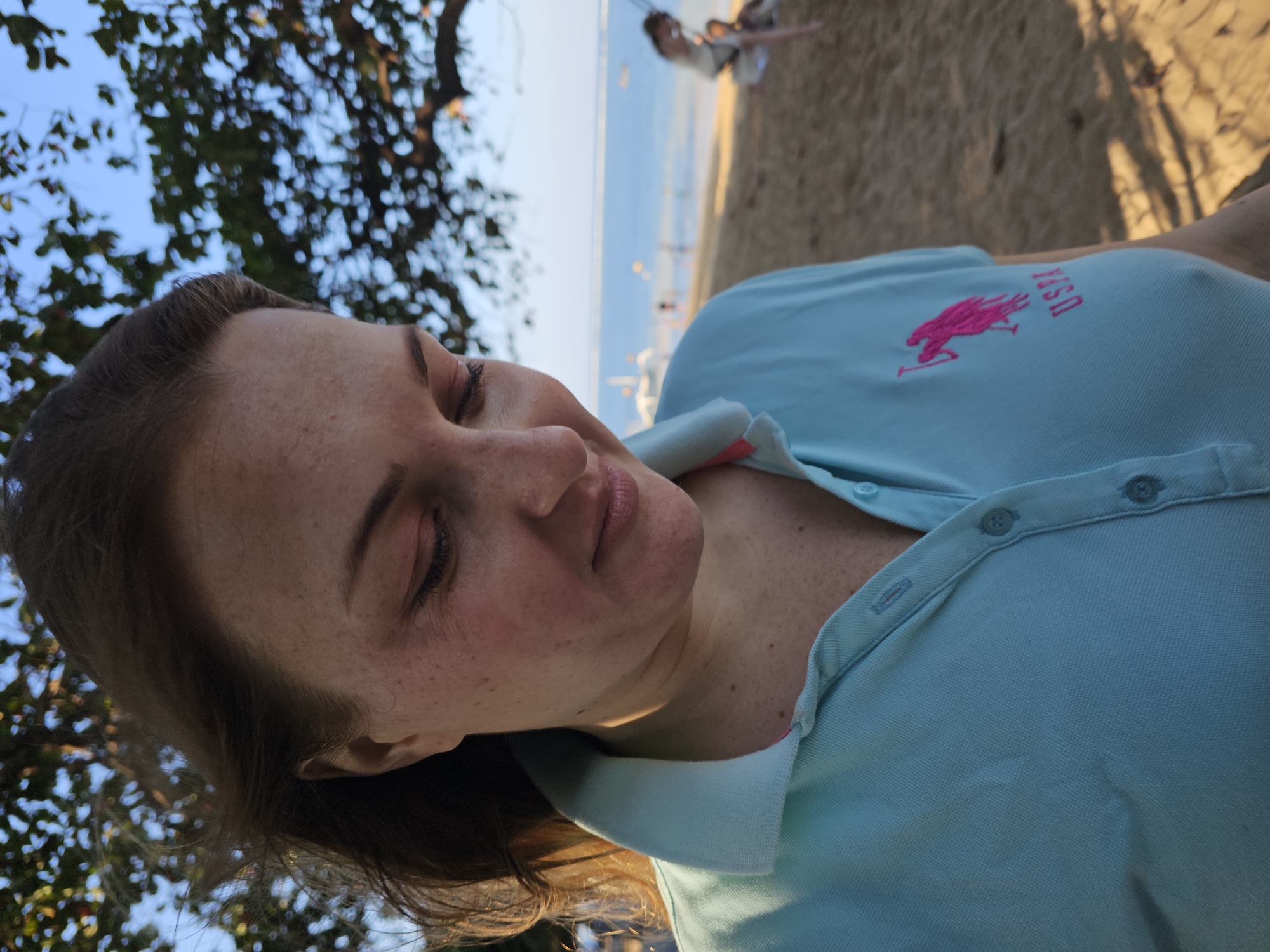 A woman with brown hair and light skin, speckled with freckles, looks down and to the right, her eyes closed and her lips slightly parted. She is wearing a light blue polo shirt with a pink horse logo and the letters "USPA" embroidered on the left side of her chest. The shirt has a collar and a button placket. The background is blurred but shows a sandy beach, the ocean, and trees with green leaves against a blue sky. In the distance, there are a few people on the beach and boats on the water. The scene appears to be a sunny day. The location is identified as Denpasar, Indonesia.