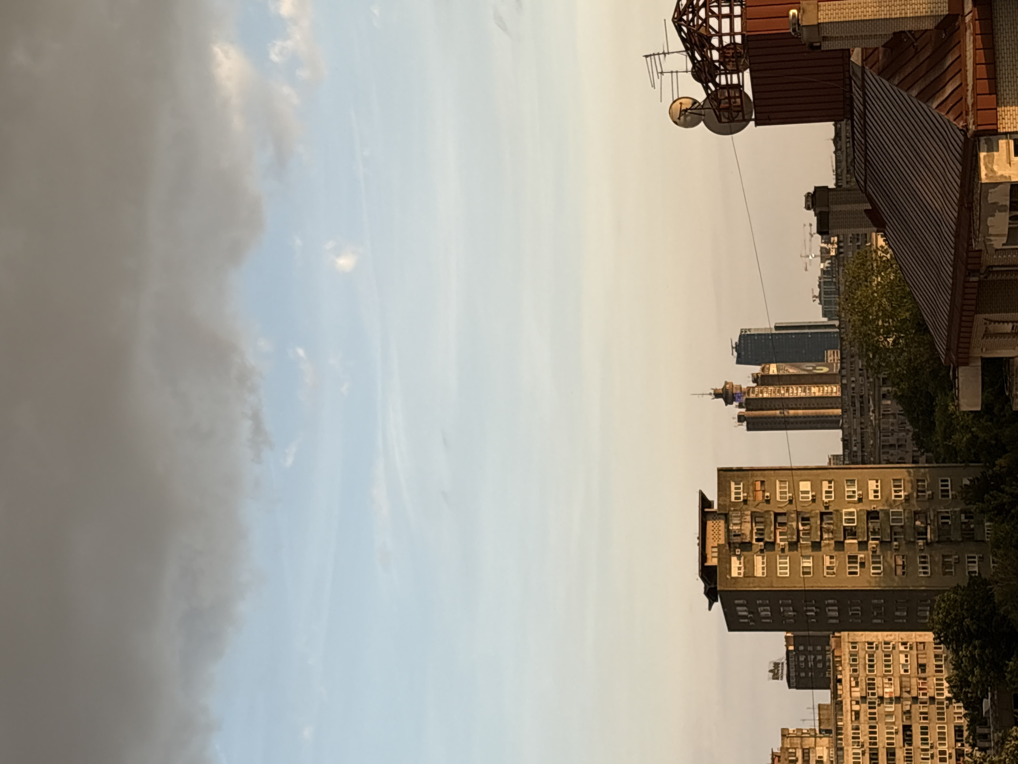 The image depicts a view of Belgrade, Serbia, from an elevated perspective. The left side is dominated by a cloudy sky. The right side shows a cityscape with various buildings, ranging from older residential structures to modern skyscrapers. In the foreground, there's a building with a rooftop antenna. The overall atmosphere appears to be during the late afternoon or early evening, with the light casting shadows and highlighting the architectural details.