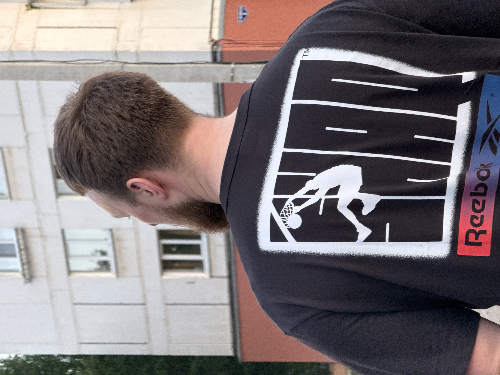 The image shows a man in Chaikovsky, Russia, from the back, wearing a black t-shirt with a large white graphic featuring a basketball player dunking a ball. The Reebok logo is displayed vertically in red and blue on the shirt. The man has short brown hair and a beard. He is standing outdoors near a multi-story building with white walls and windows. A red building is partially visible in the background as well.